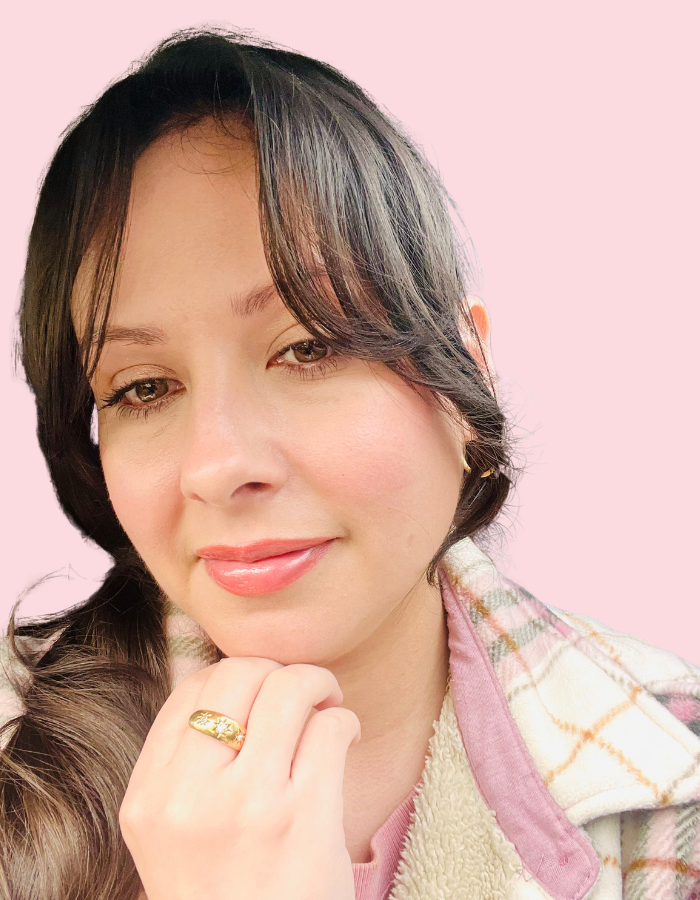 A close-up portrait of Dani with dark hair wearing a gold ring, set against a solid light pink background.