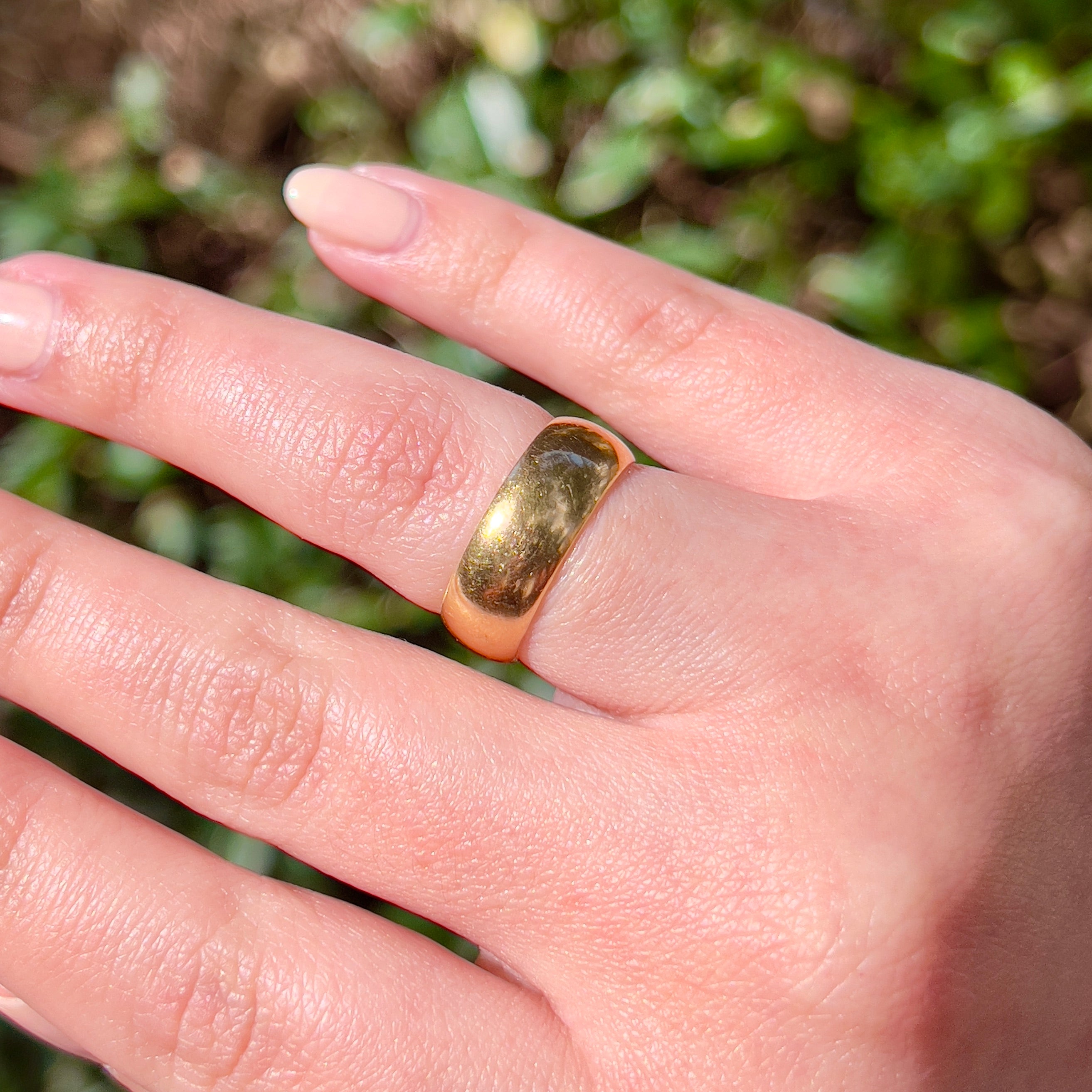 A close-up of a hand wearing a gold band ring outdoors, with blurred green foliage in the background.