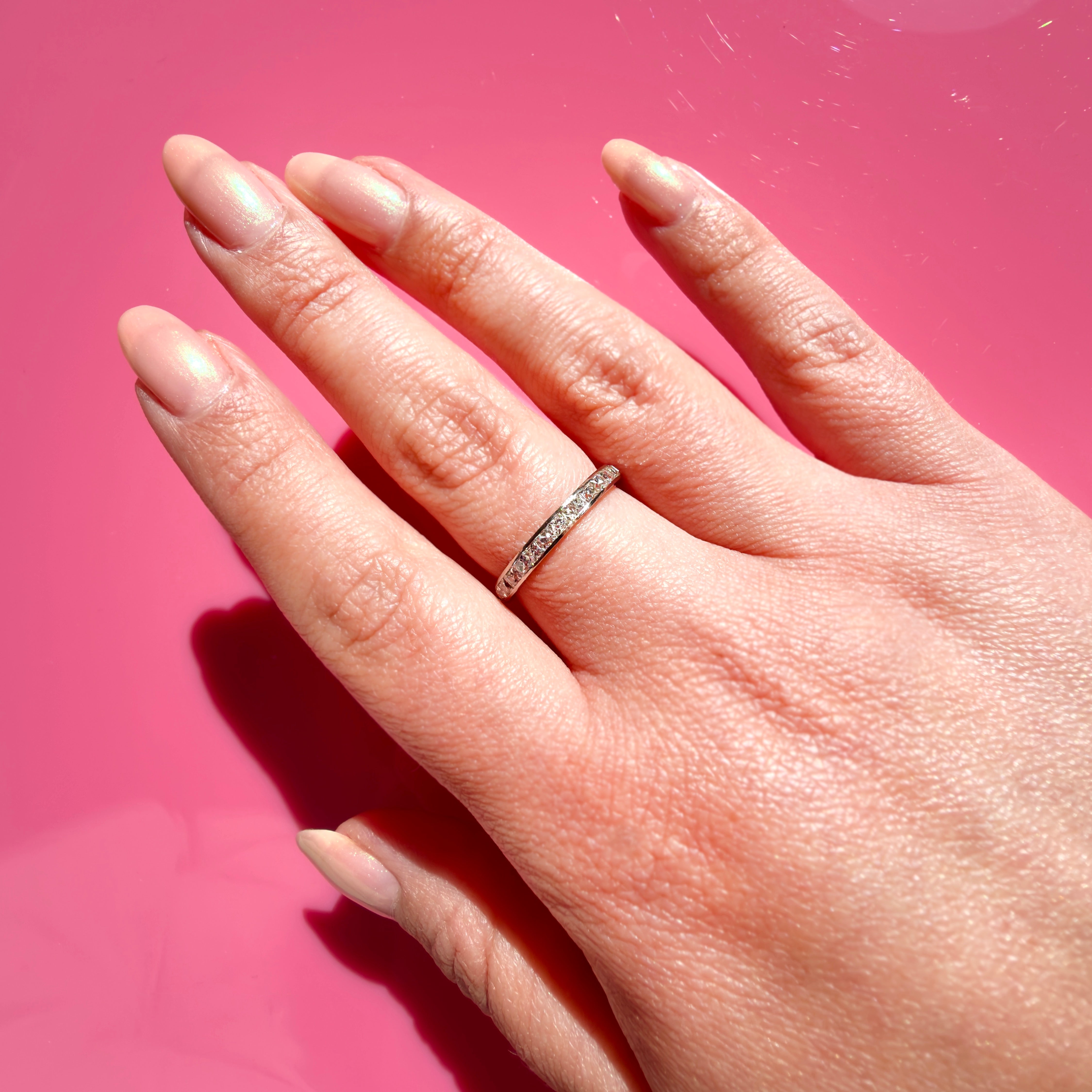 A platinum band ring with small French cut diamonds set along the top, worn on a person's ring finger against a solid pink background.