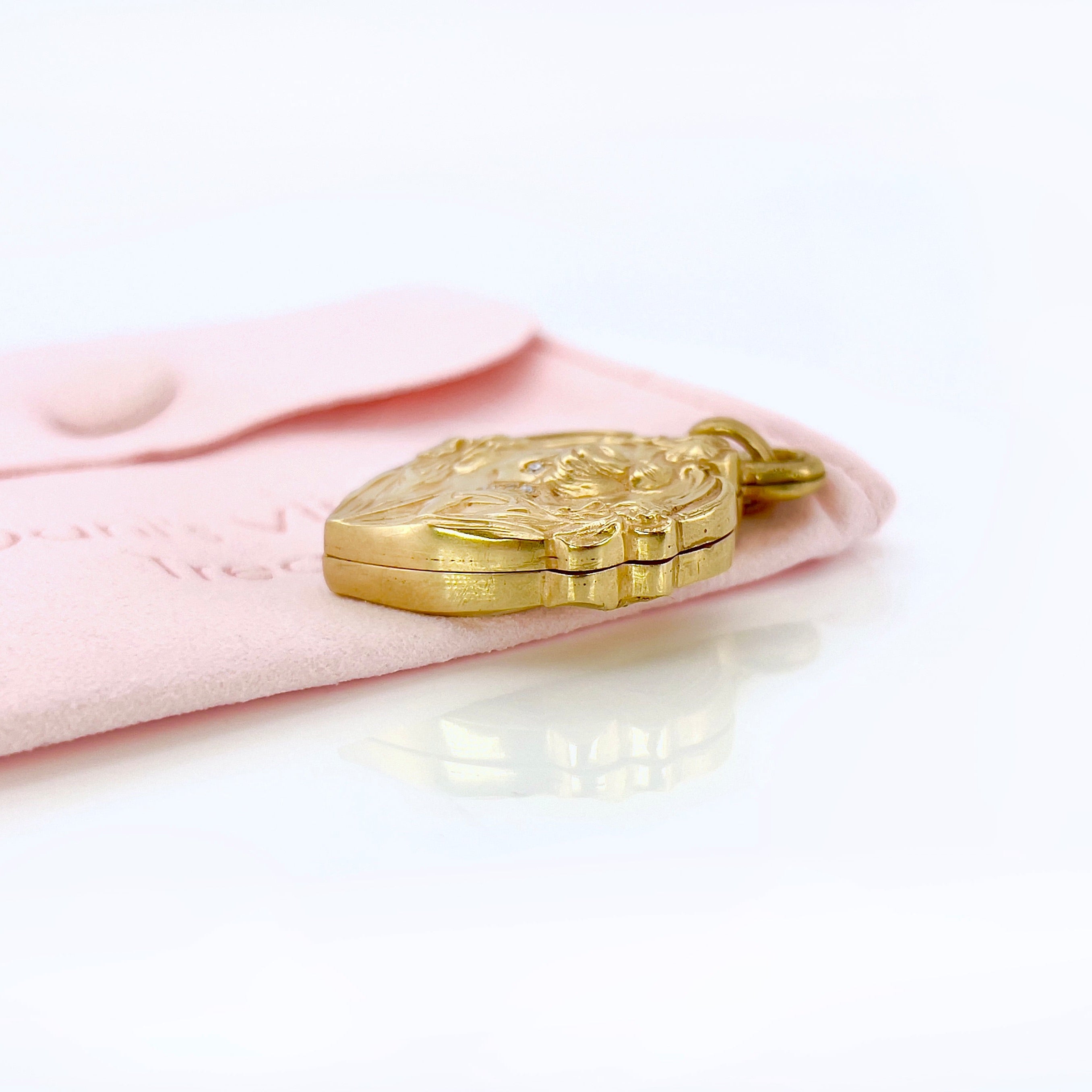 An Art Nouveau heart-shaped locket with gold detailing, featuring a central figure with a bun and flowers on the front, on a pink pouch.