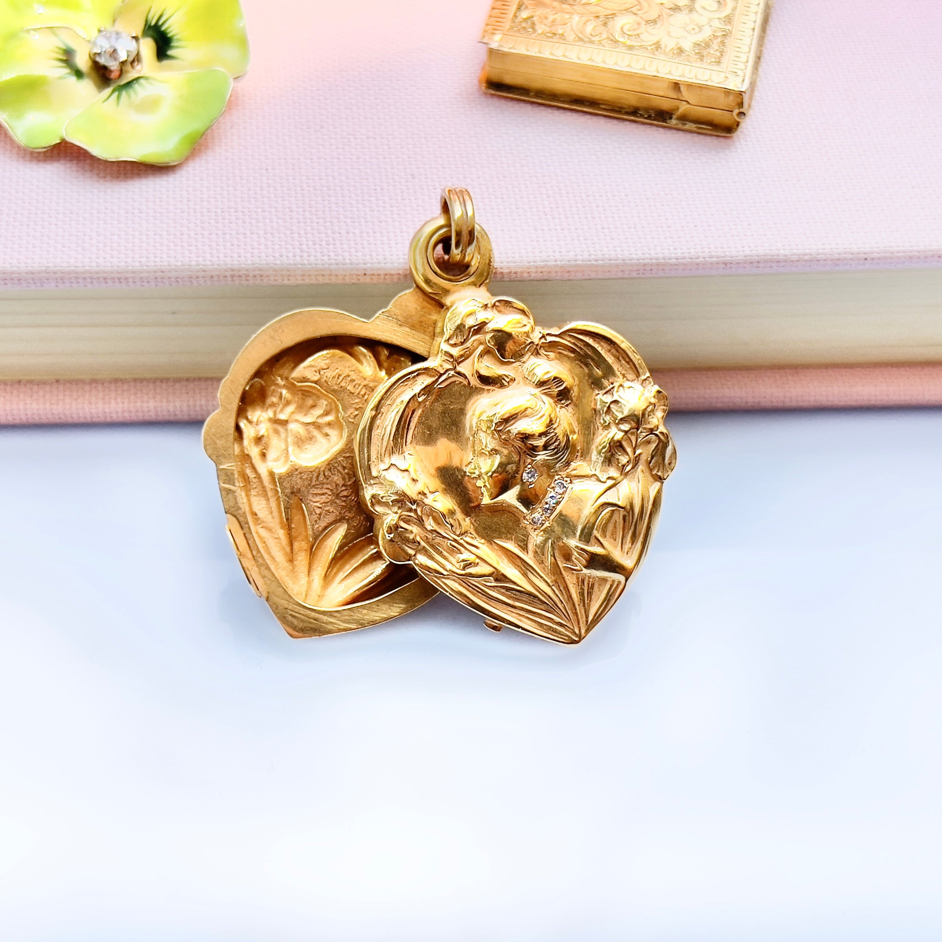 An Art Nouveau heart-shaped locket with gold detailing, featuring a central figure with a bun and flowers on the front, on a blurred background.