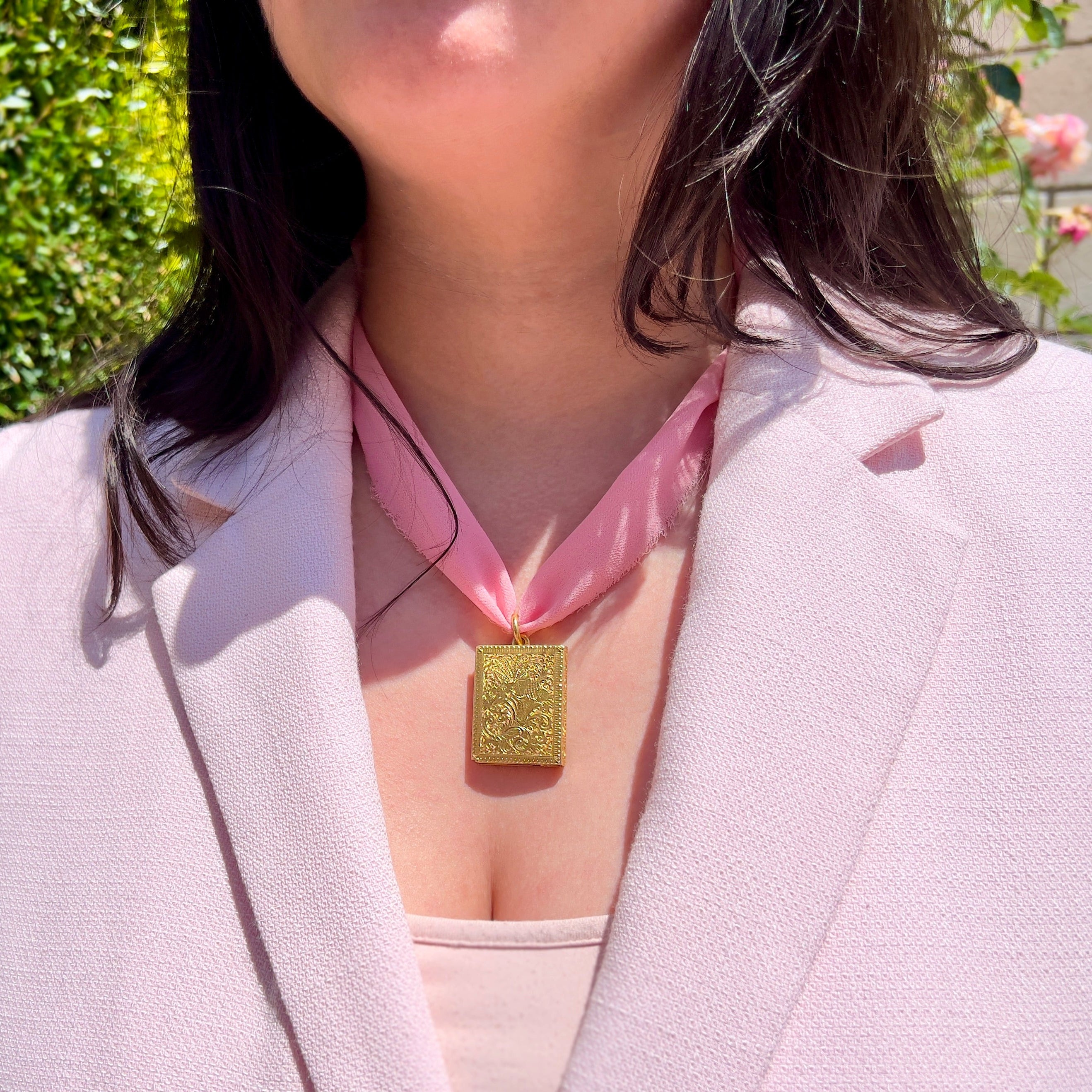 A close up of a person wearing a gold victorian book locket with intricate design on a pink ribbon against an outdoor background