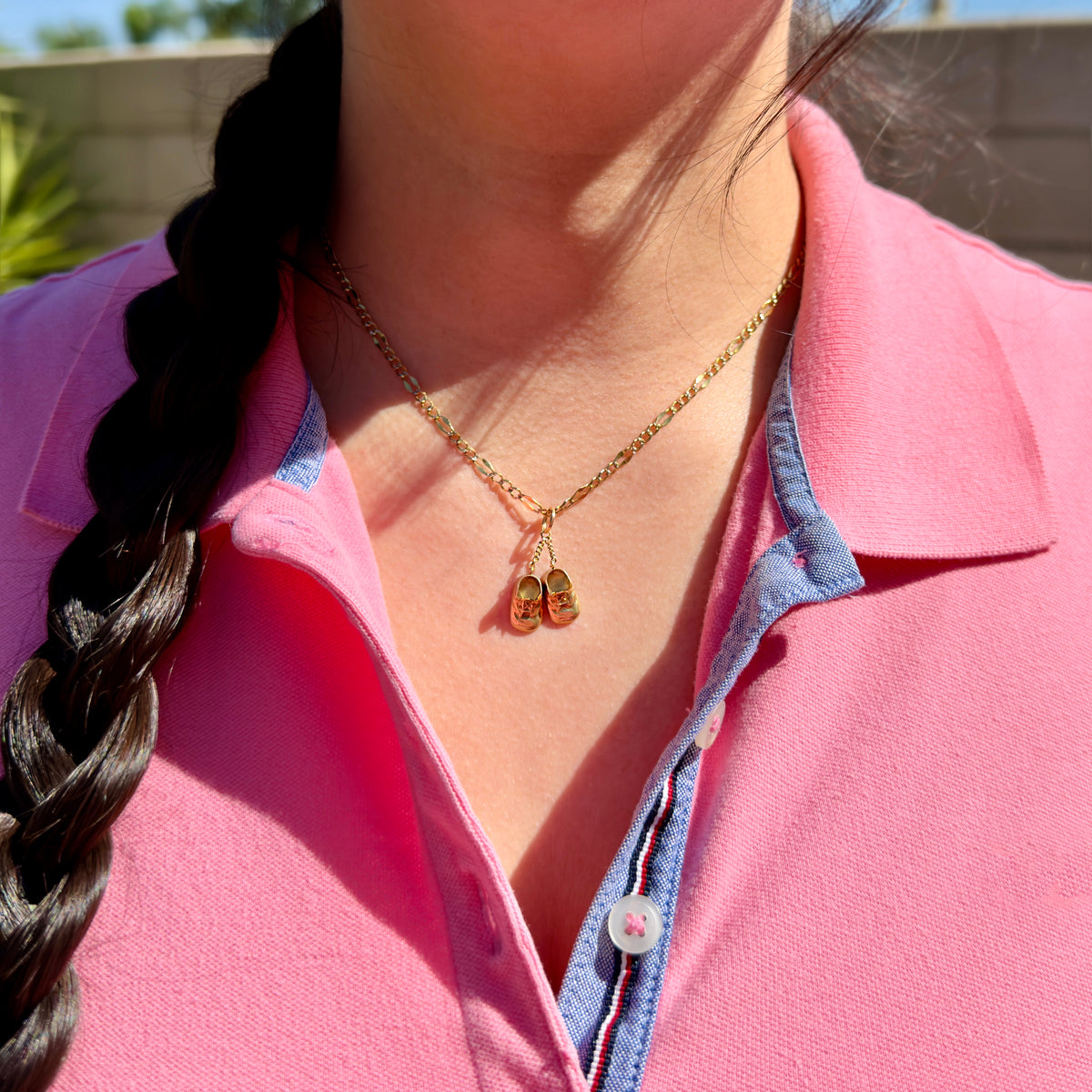 A close-up of a person wearing a delicate gold necklace with a baby shoes charm, styled with a pink polo shirt and blue collar trim.