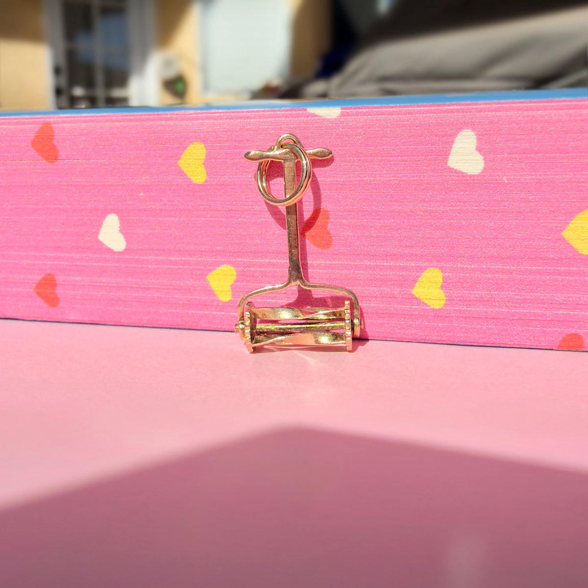 A hand lawn mower charm with a loop at the top, photographed standing upright against a bright pink background with scattered heart shapes.