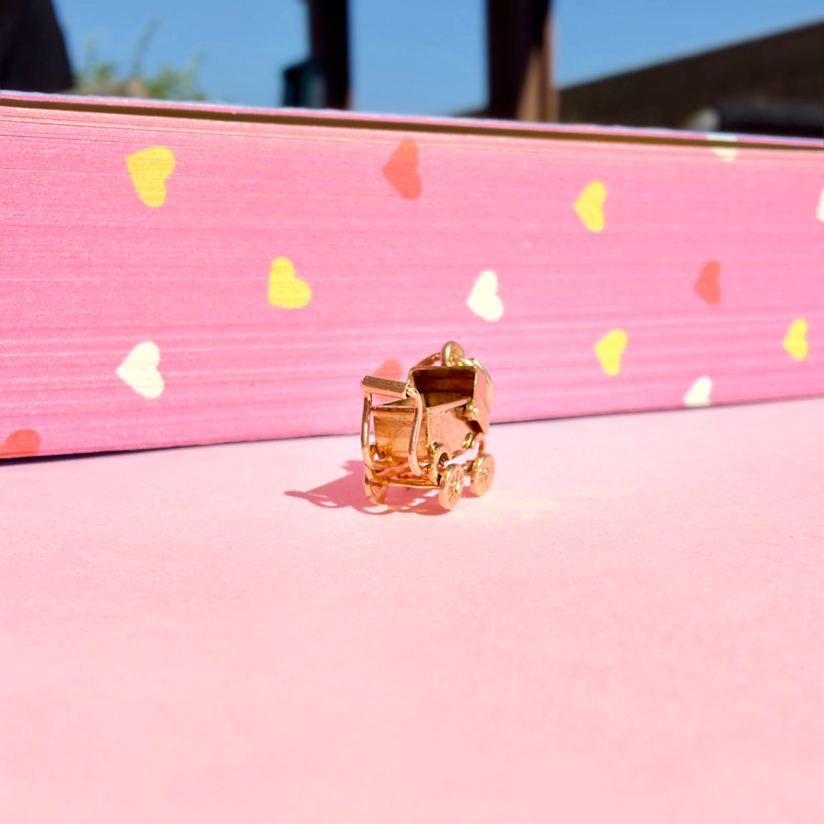 A small gold miniature stroller charm, resting on a pink surface in front of a pink book decorated with colorful hearts.