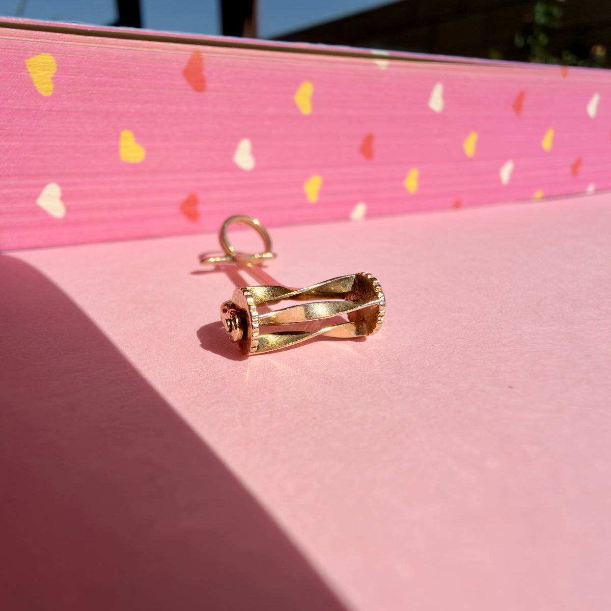 A hand lawn mower charm with a loop at the top, photographed laying down against a bright pink background with scattered heart shapes.
