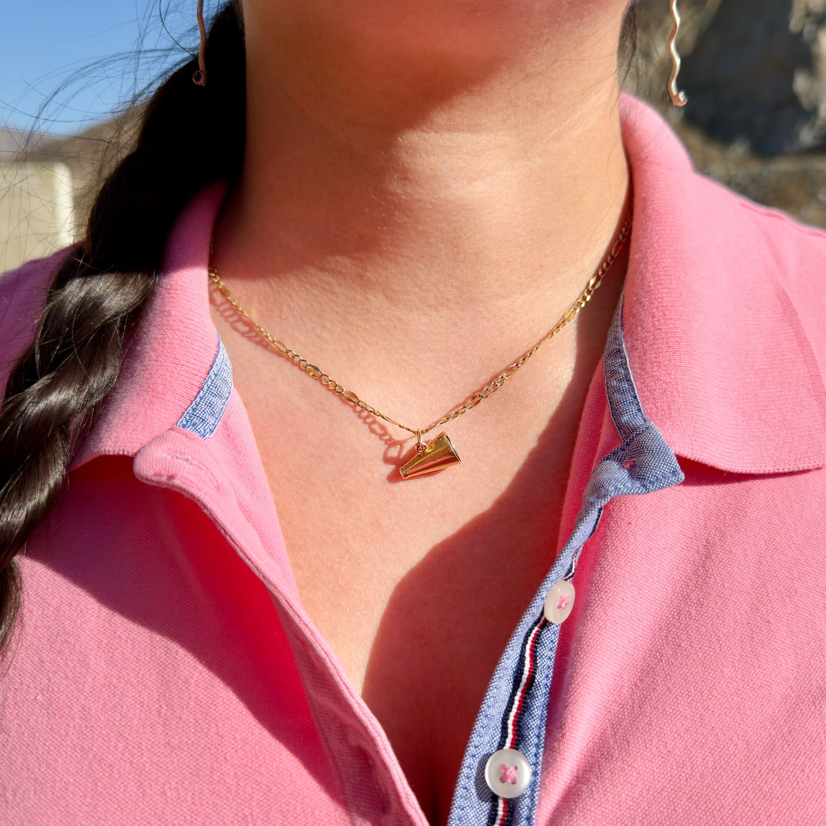 A close-up of a model wearing a delicate gold chain necklace with a small megaphone charm, styled with a pink polo shirt.