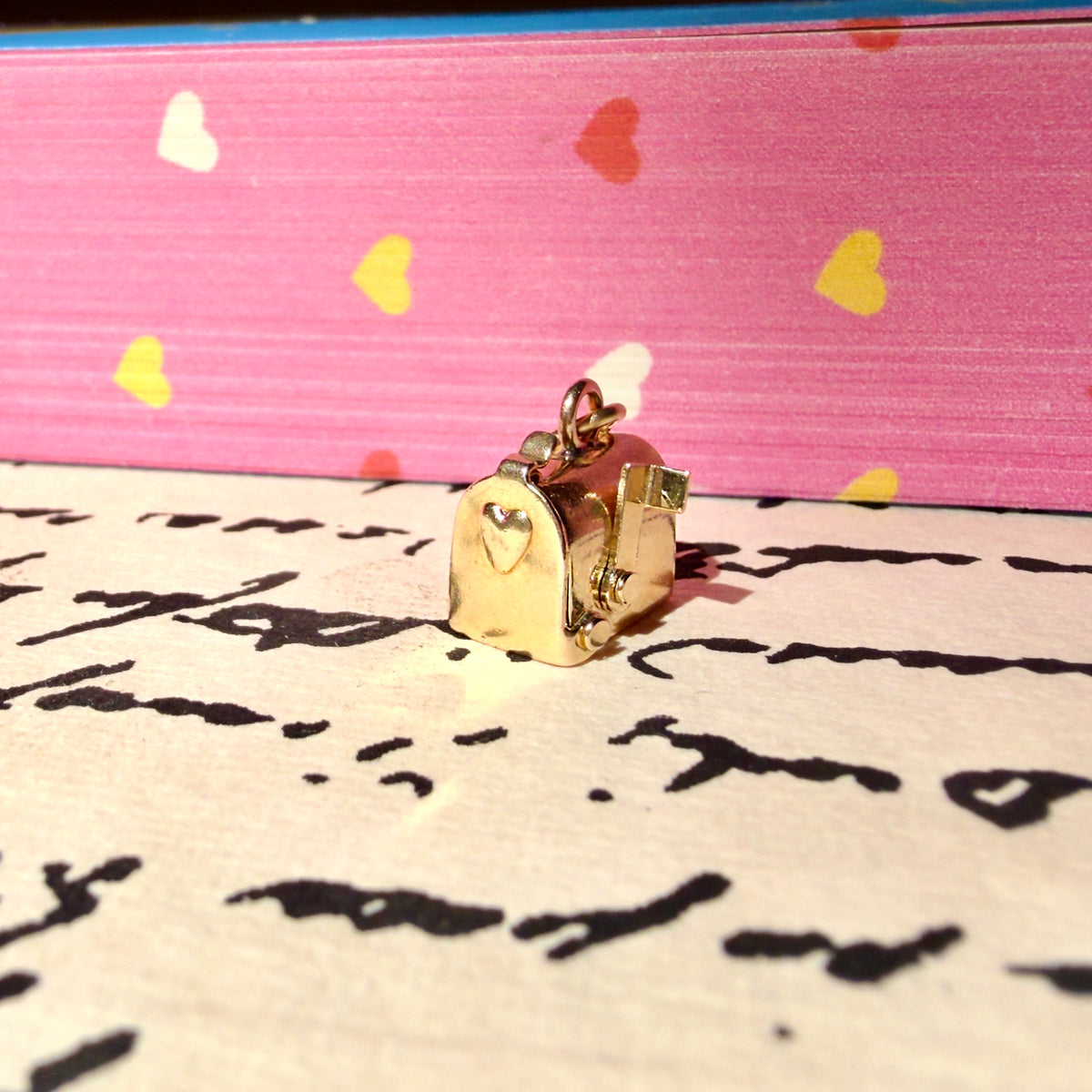 A small gold-toned miniature mailbox charm with a heart detail, resting on a piece of paper with cursive writing, with a pink background featuring heart patterns.