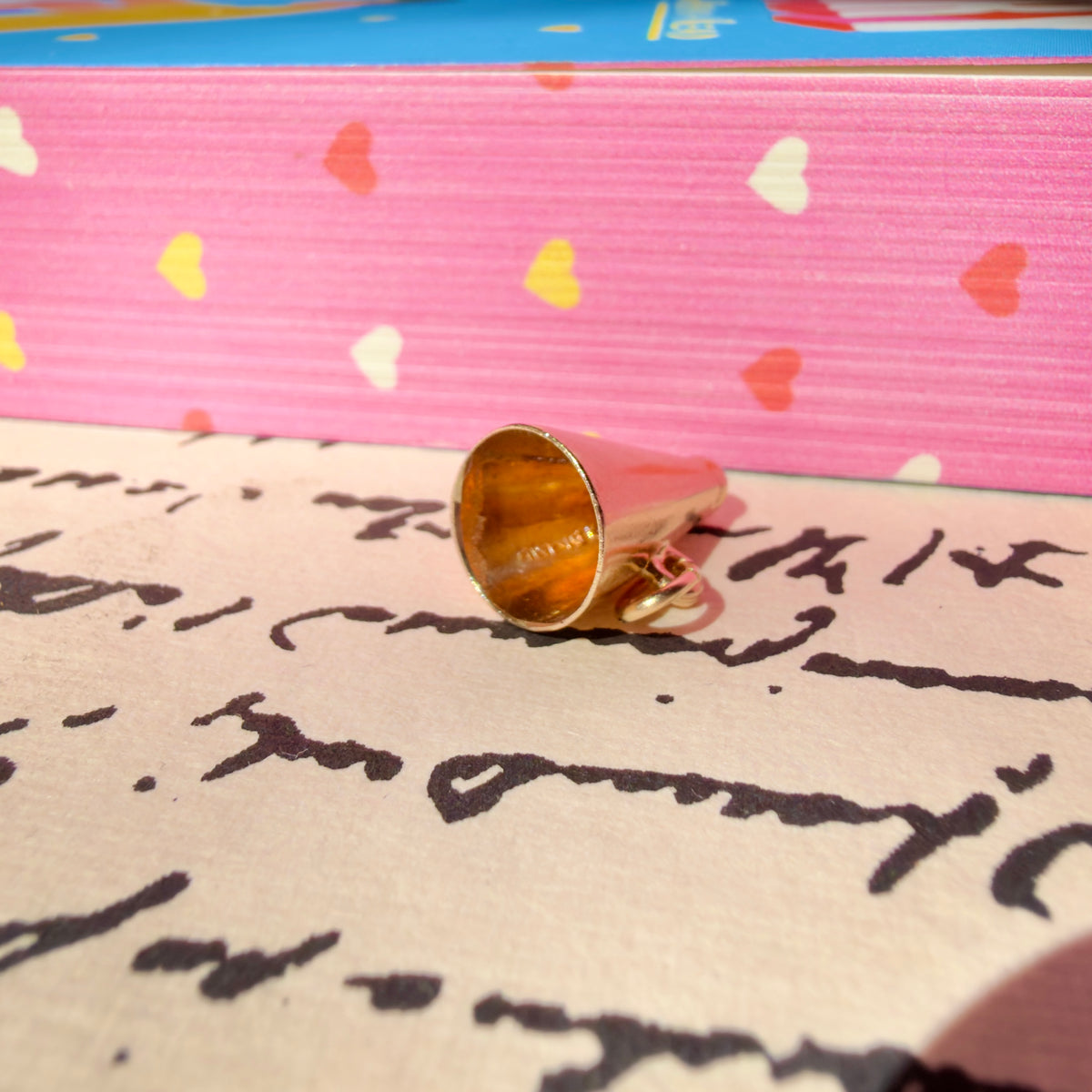 A gold megaphone charm lies on a piece of paper with cursive writing, with a pink patterned box in the background.