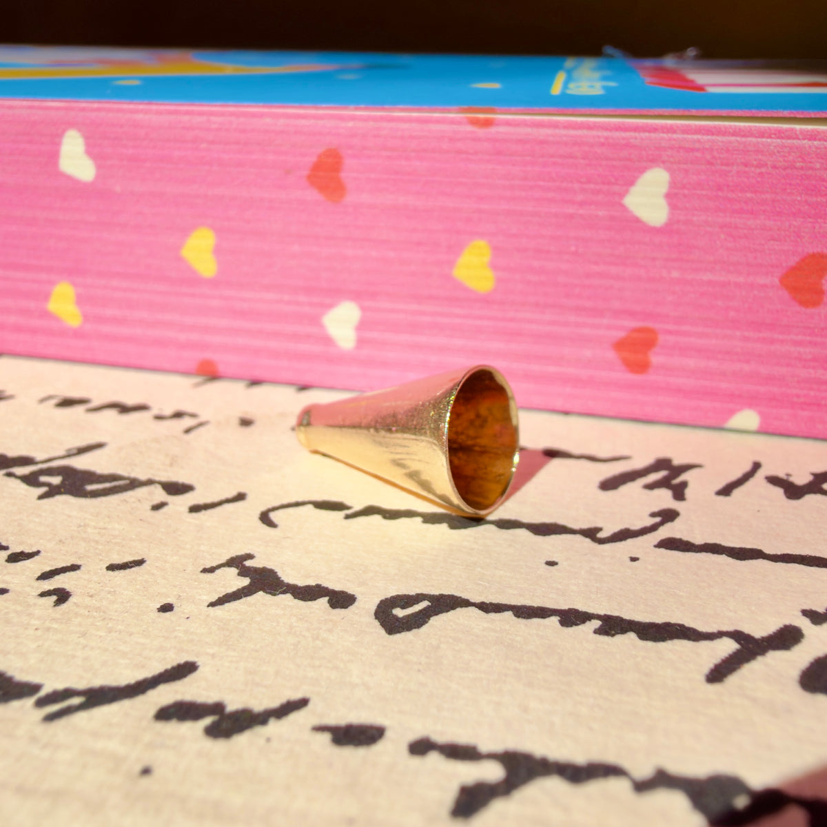 A small metallic gold megaphone charm, resting on a piece of cream-colored paper with cursive writing. In the background, a pink book with colorful heart patterns is visible.
