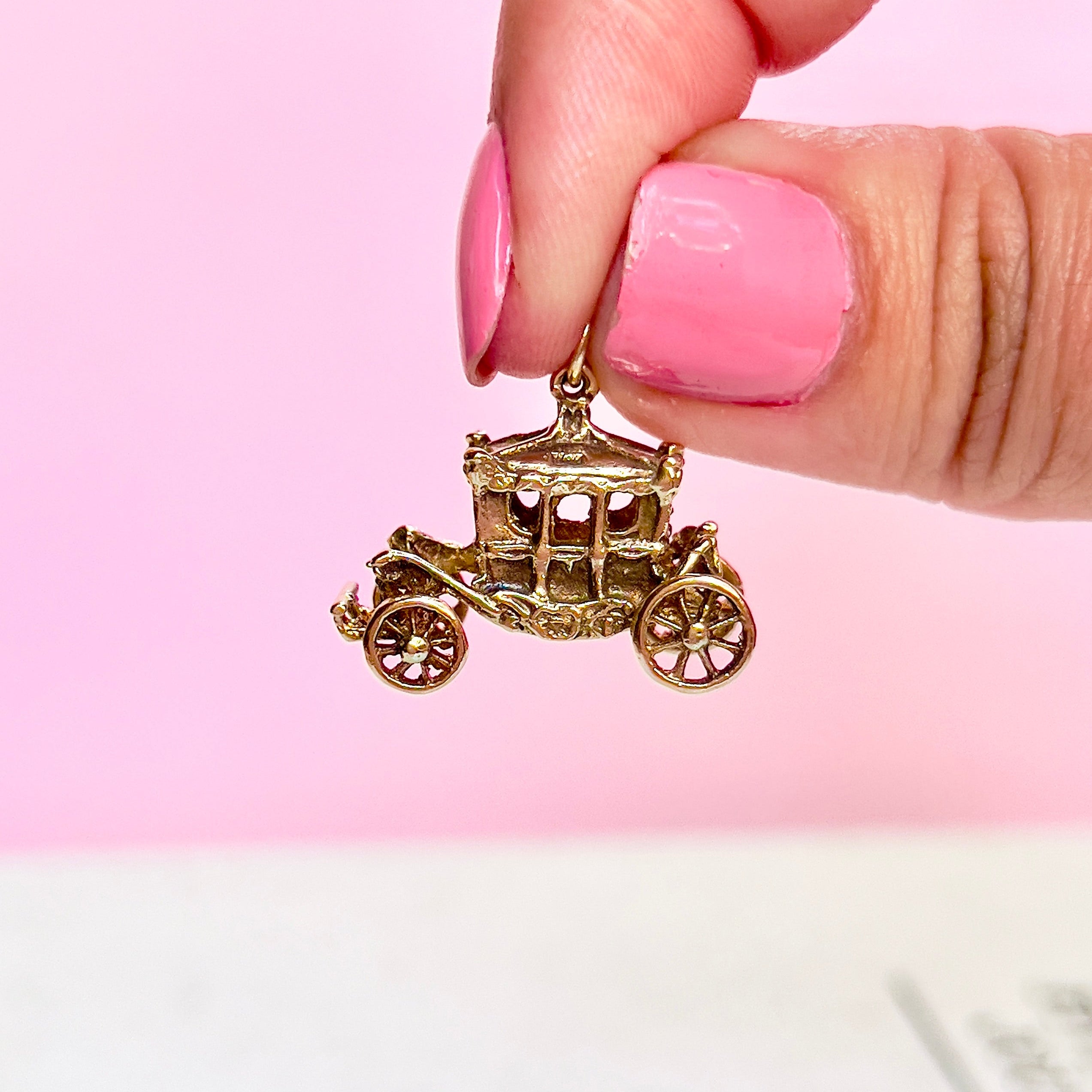 A vintage 9KT gold charm shaped like a Cinderella carriage with moving wheels, held by a hand with a pink background.