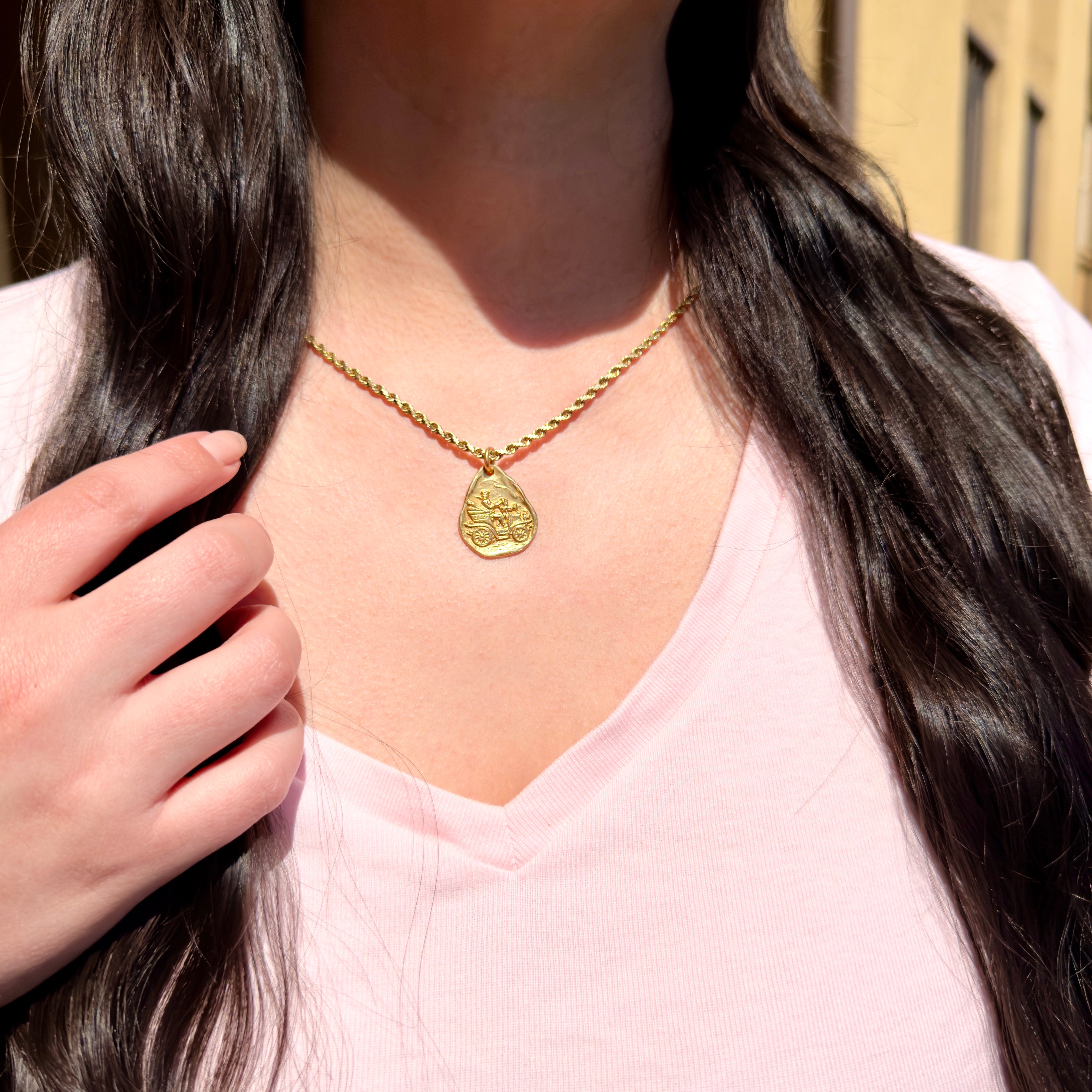 Person wearing a gold necklace with a pendant, outdoors.