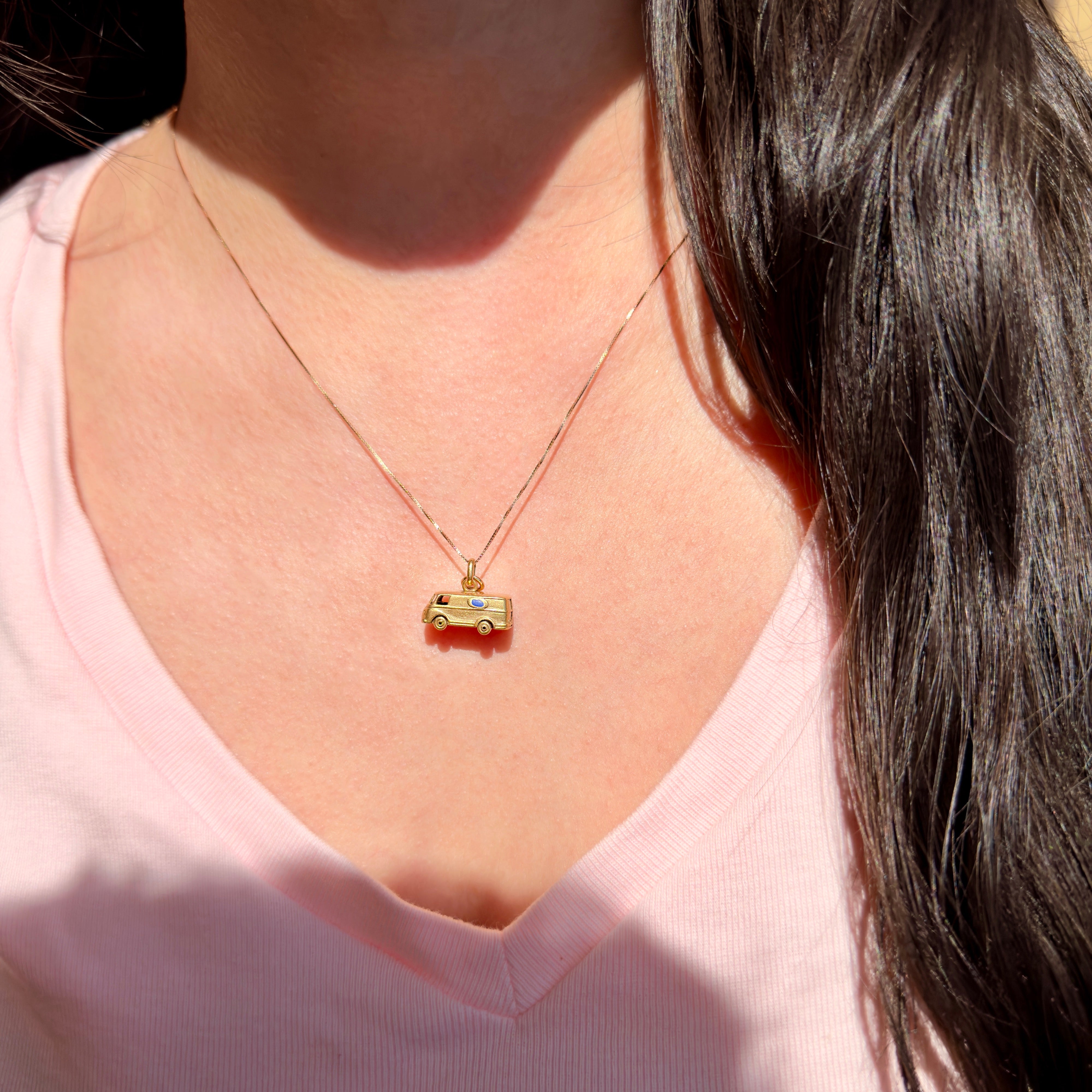 Person wearing a gold necklace with a small 1960s van charm, set against a blurred background.