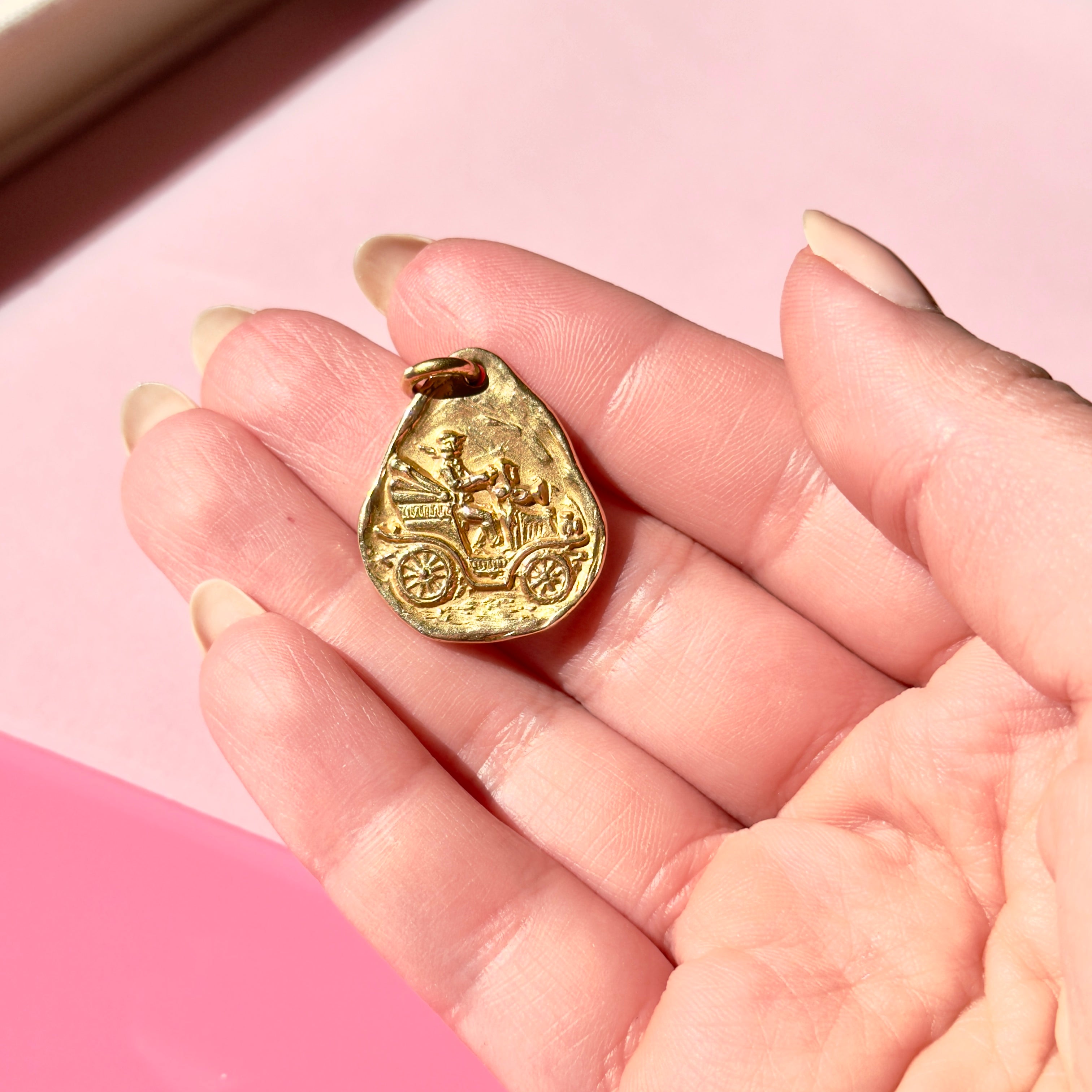 Gold pendant with a car design held in a hand against a pink background