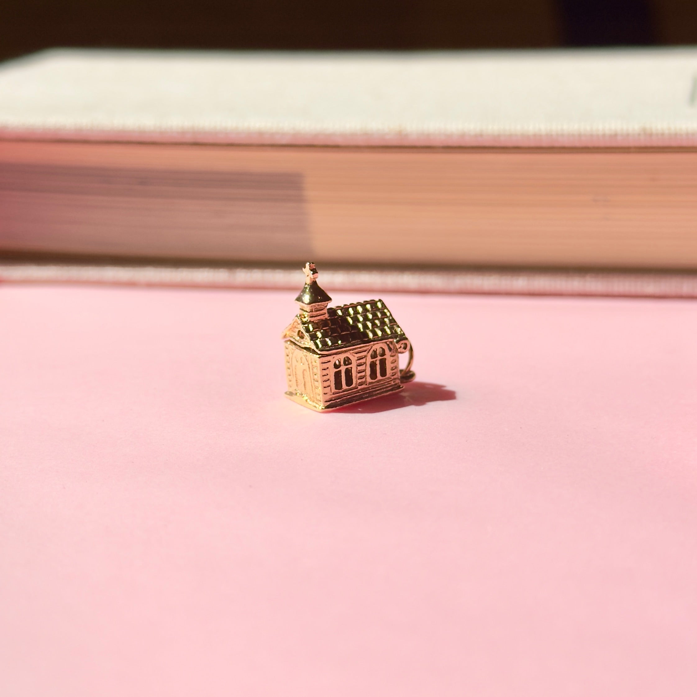 Small vintage gold church charm on a pink surface with a book in the background