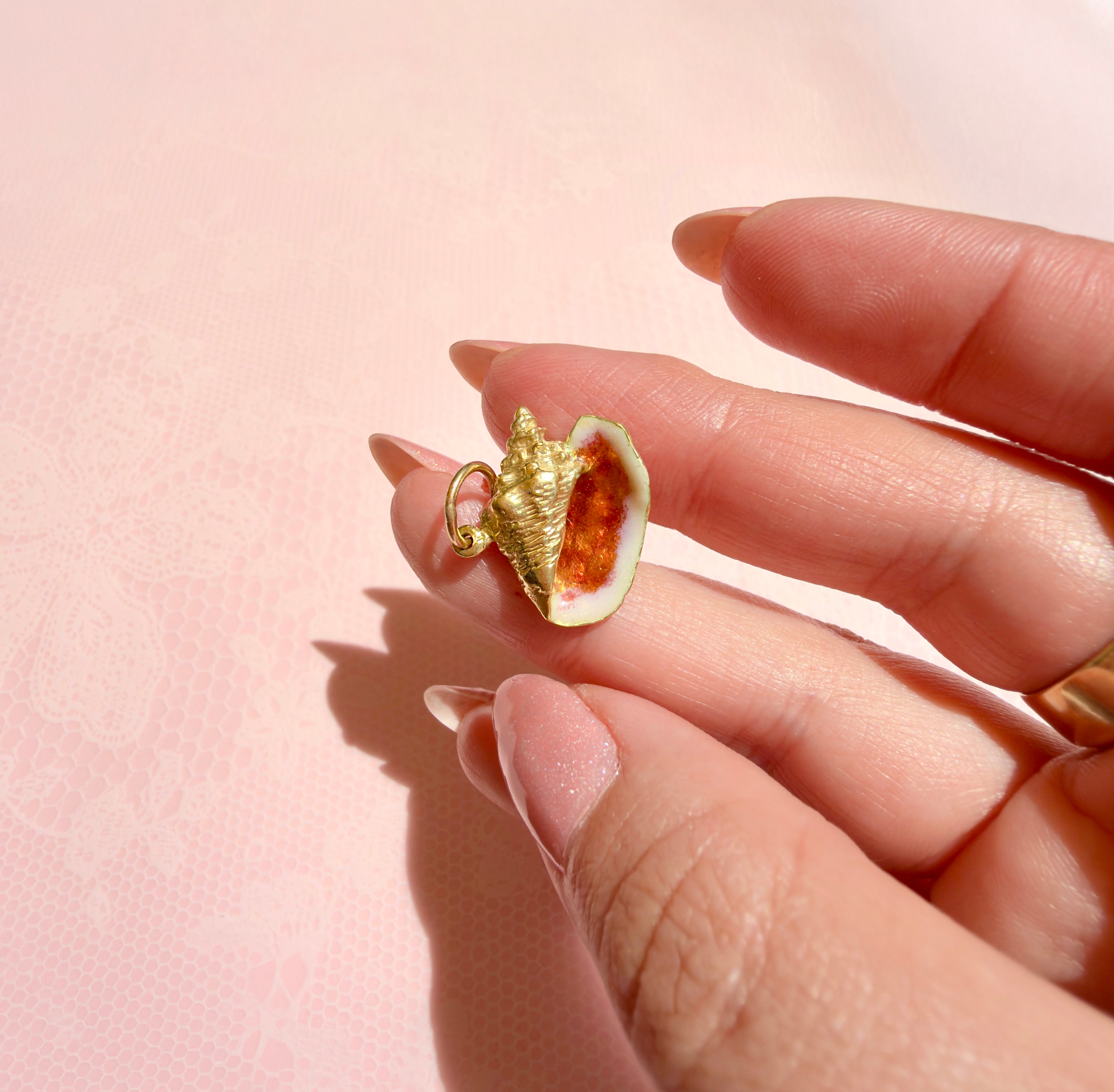 A hand holding an 18K yellow gold vintage conch shell charm with orange enamel against a pink background.