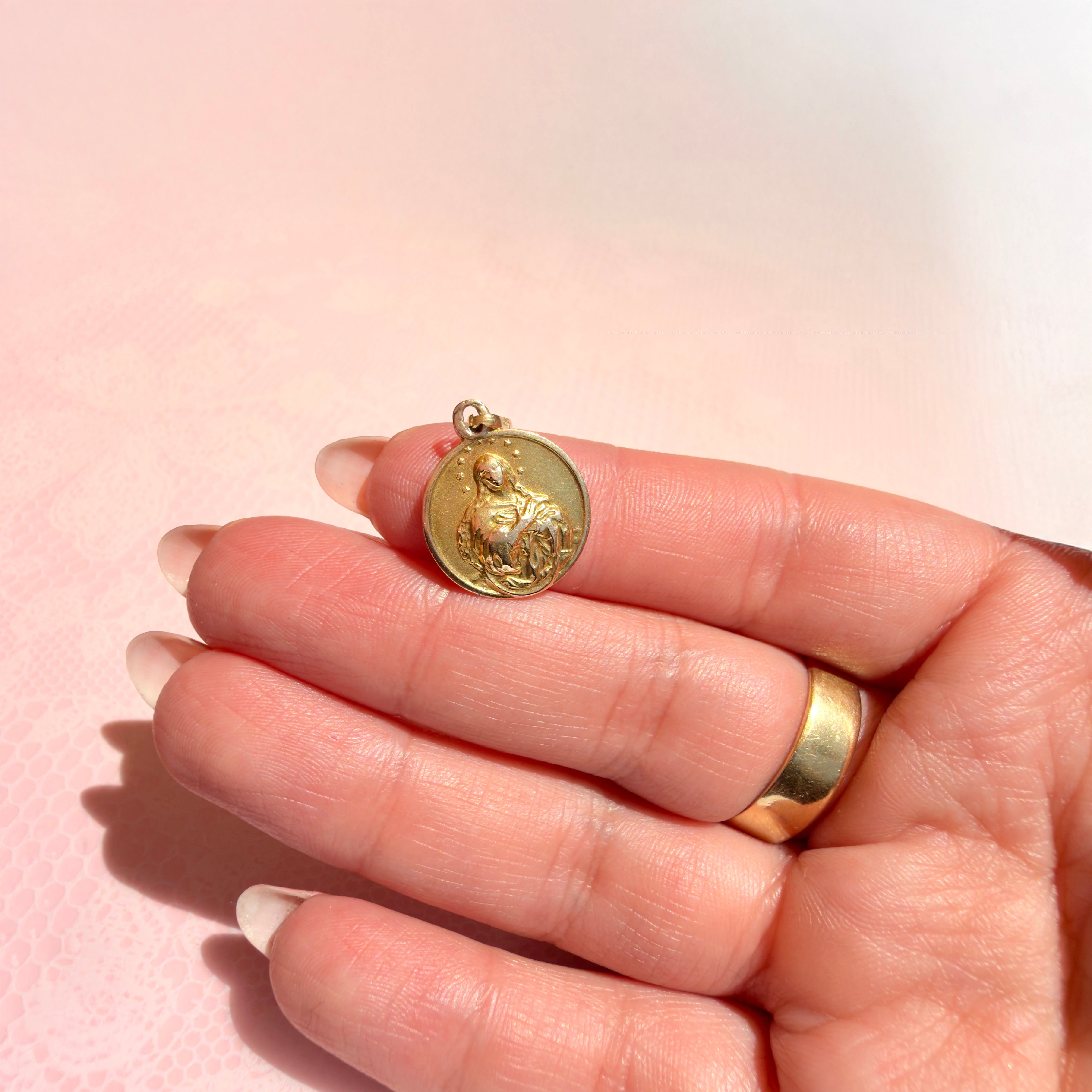A hand holding a gold pendant with a religious figure on a pink background
