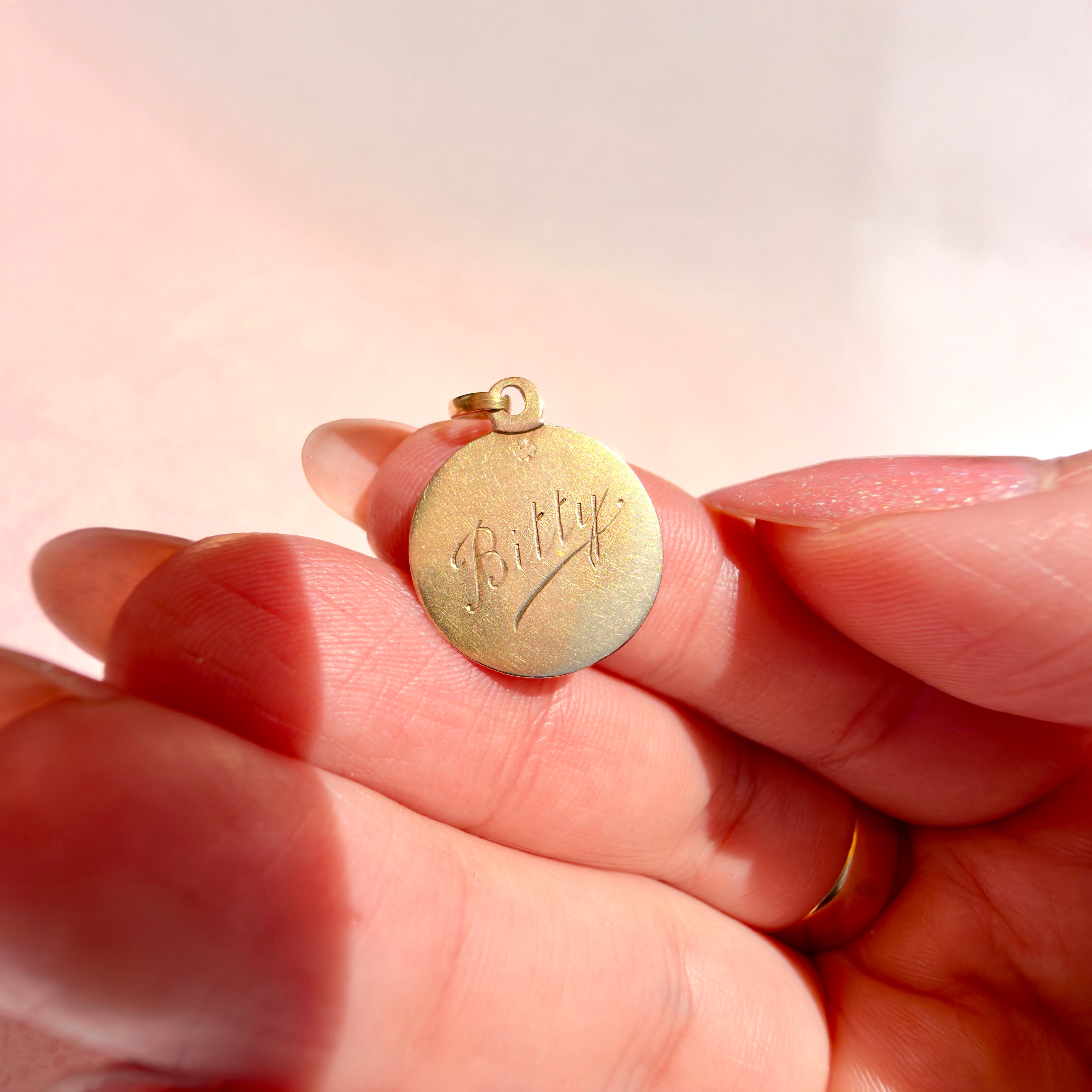 A hand holding a gold pendant with "Bitty" on a pink background