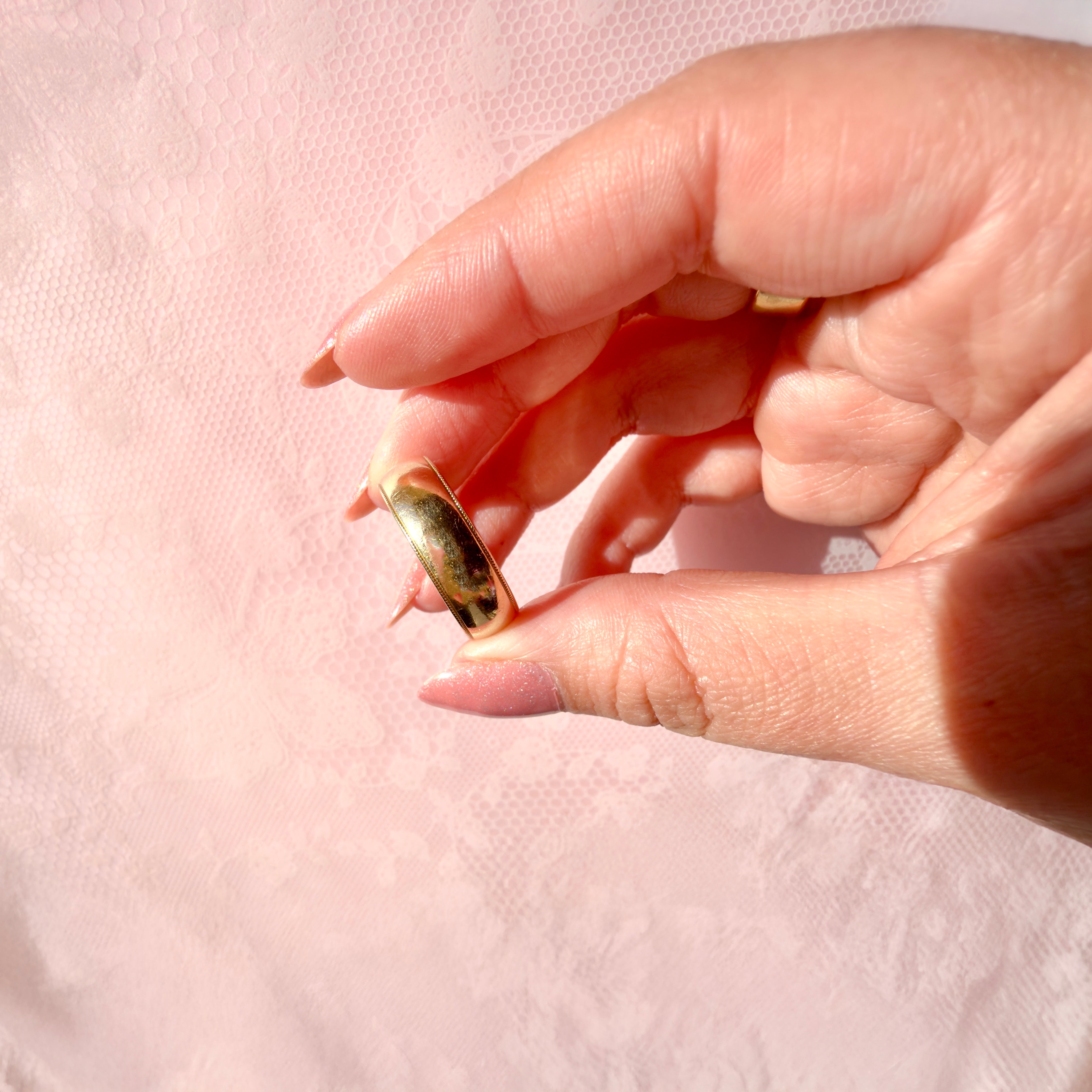 Close-up of a hand holding a gold ring against a light pink background