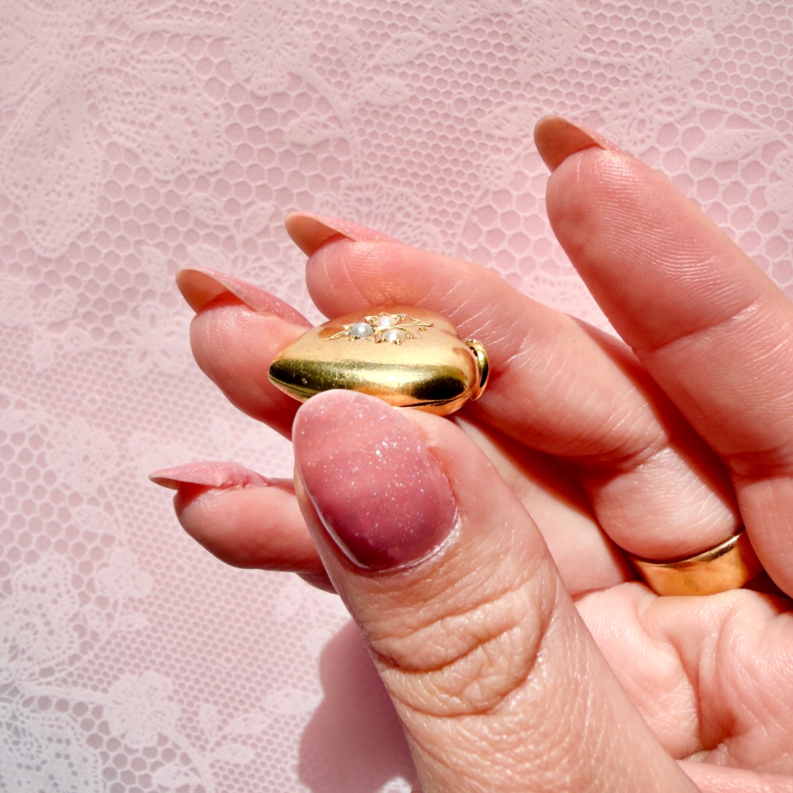 Hand holding a gold heart-shaped pendant with pearls on a pink background