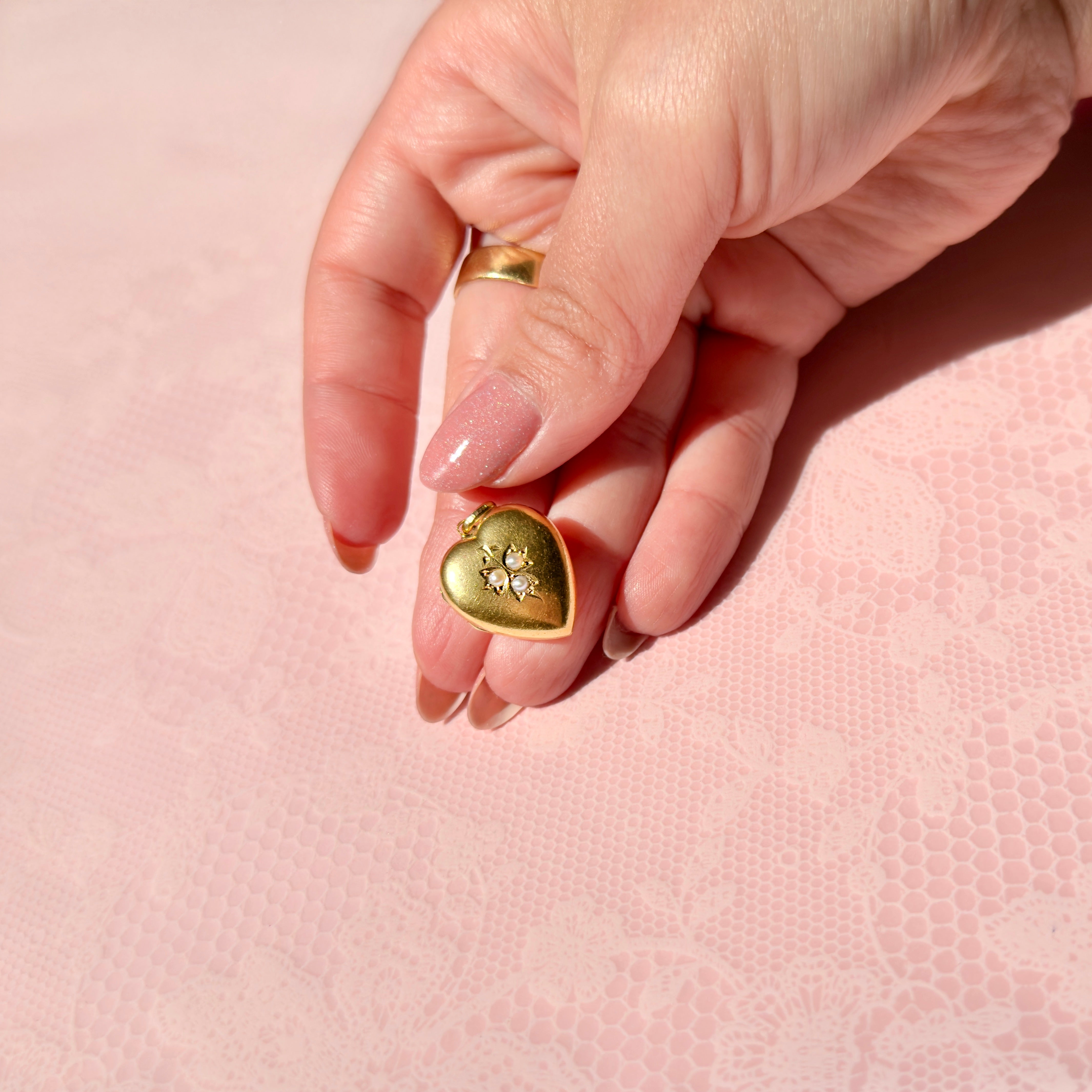 Hand holding a gold heart-shaped pendant with pearls on a pink background