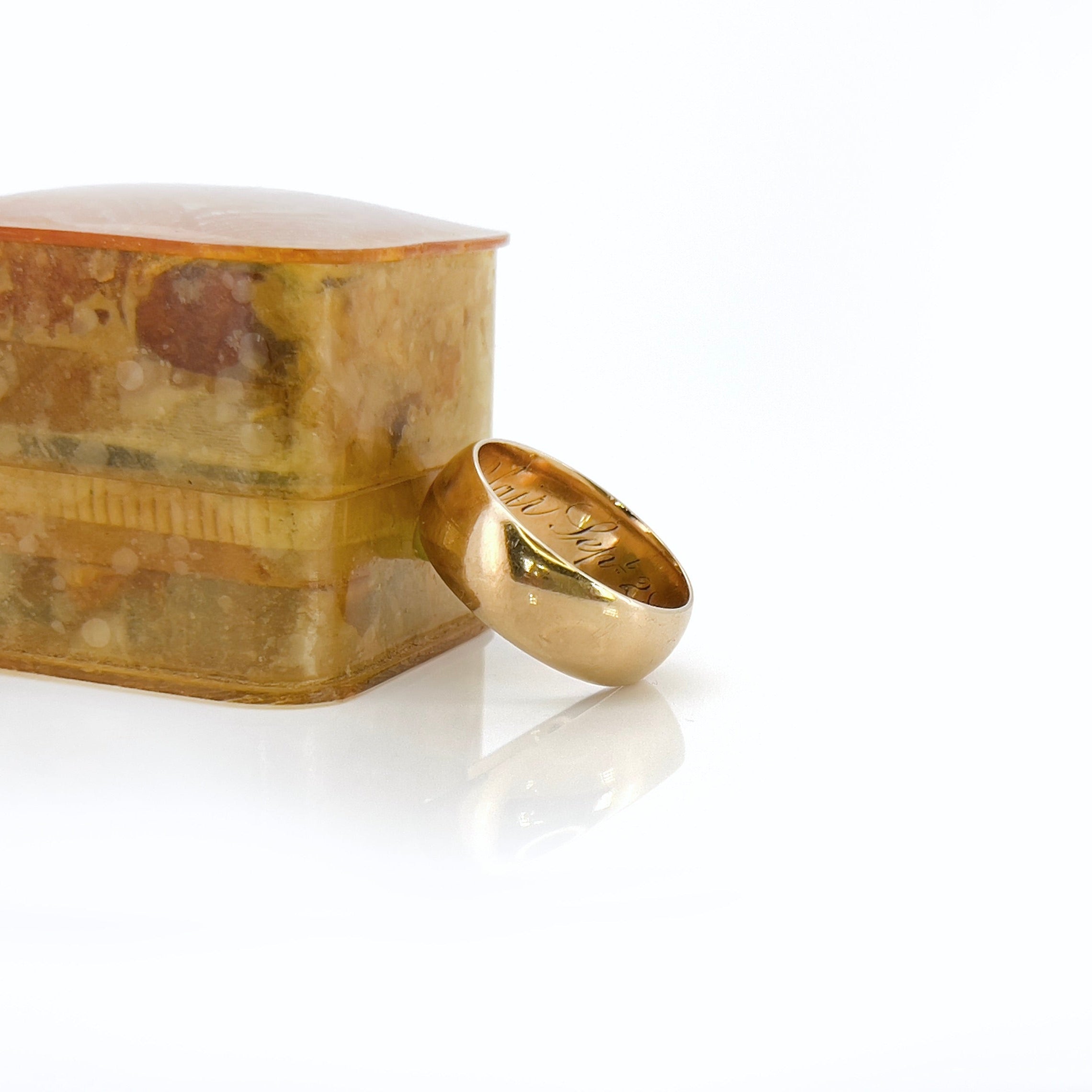 A Victorian gold band with a patina and inscription leaning on a light brown vintage ring box in front of a white background.