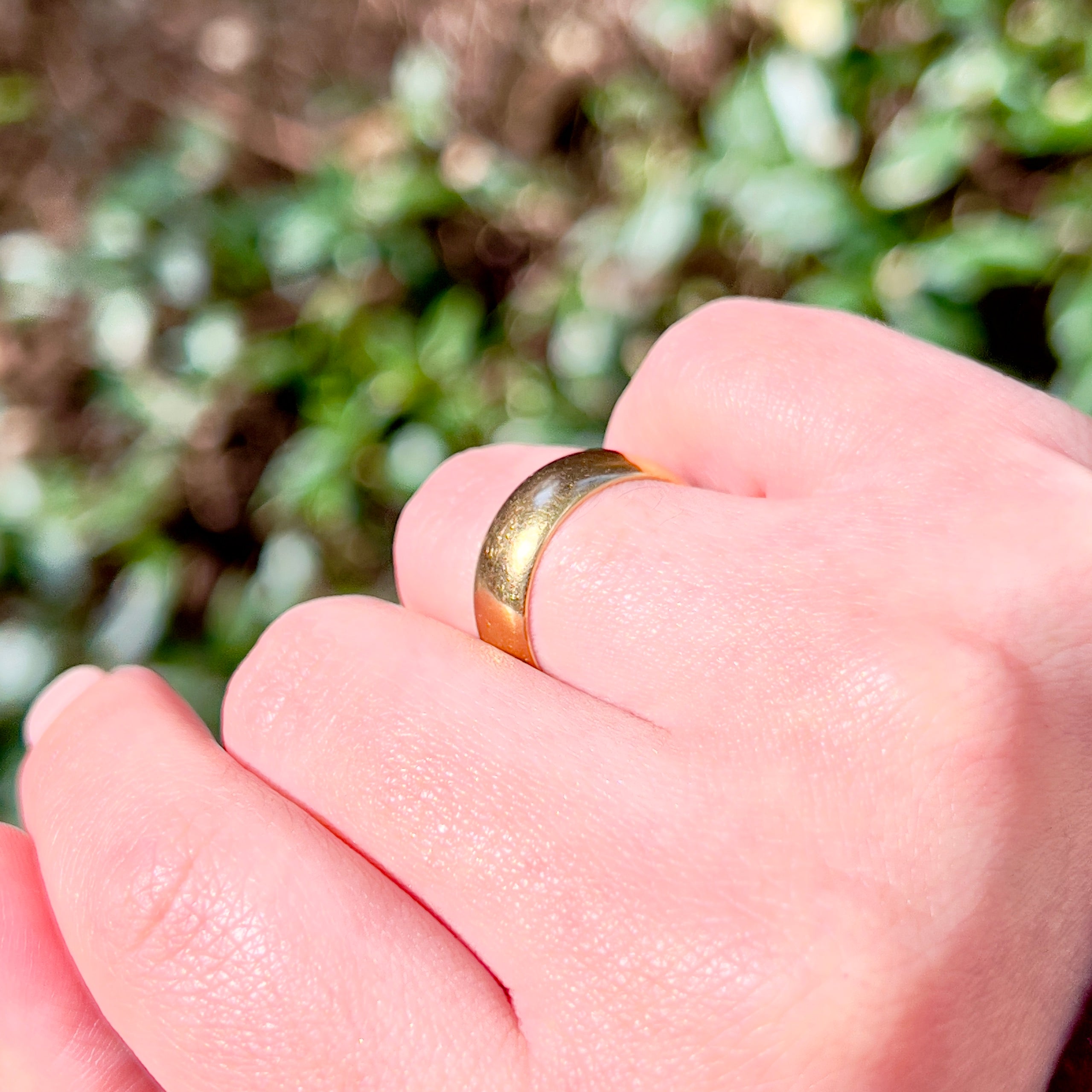 A close-up of a hand wearing a simple gold band ring, with a soft-focus background of green foliage.