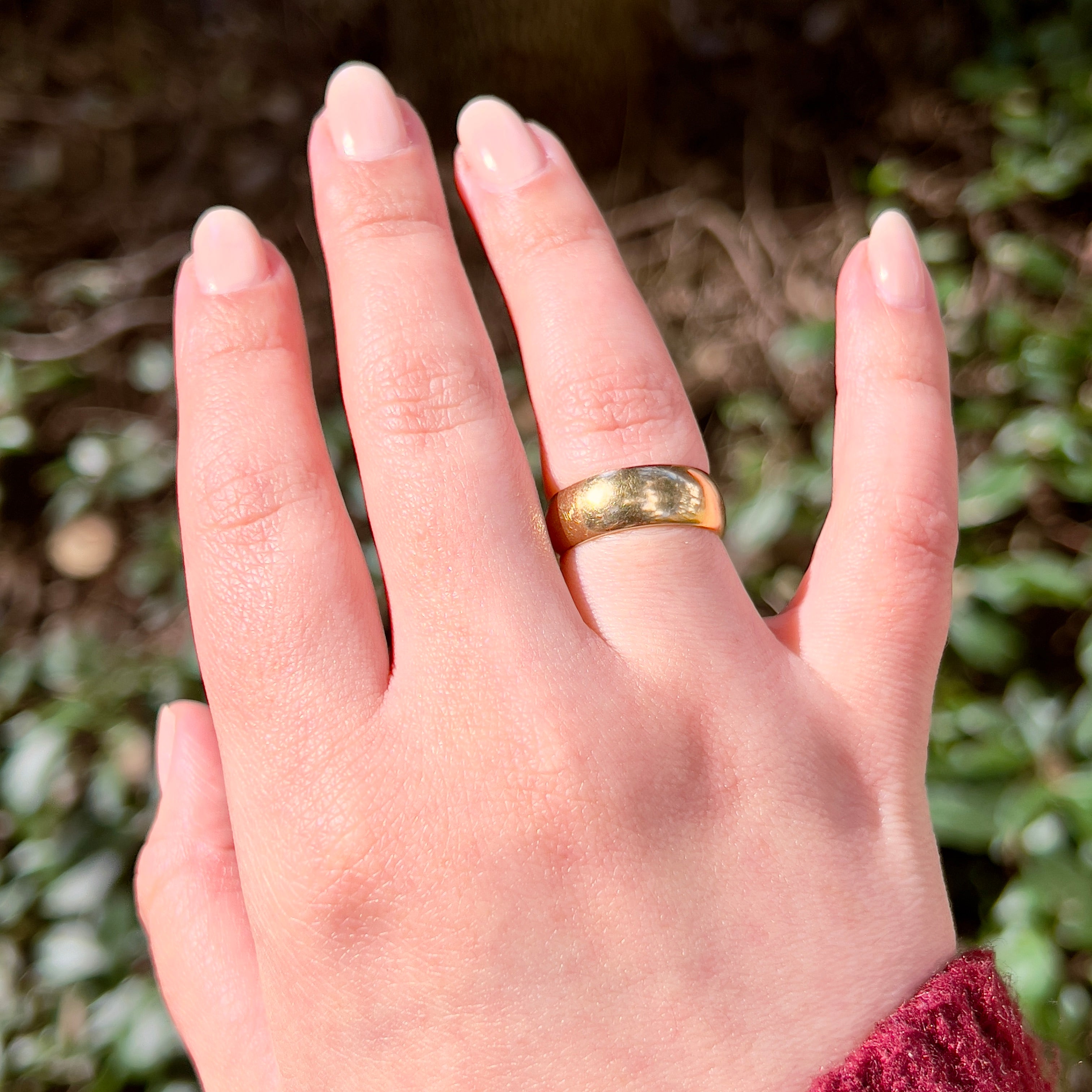 A close-up of a hand wearing a simple gold band ring, photographed outdoors with blurred green foliage in the background.