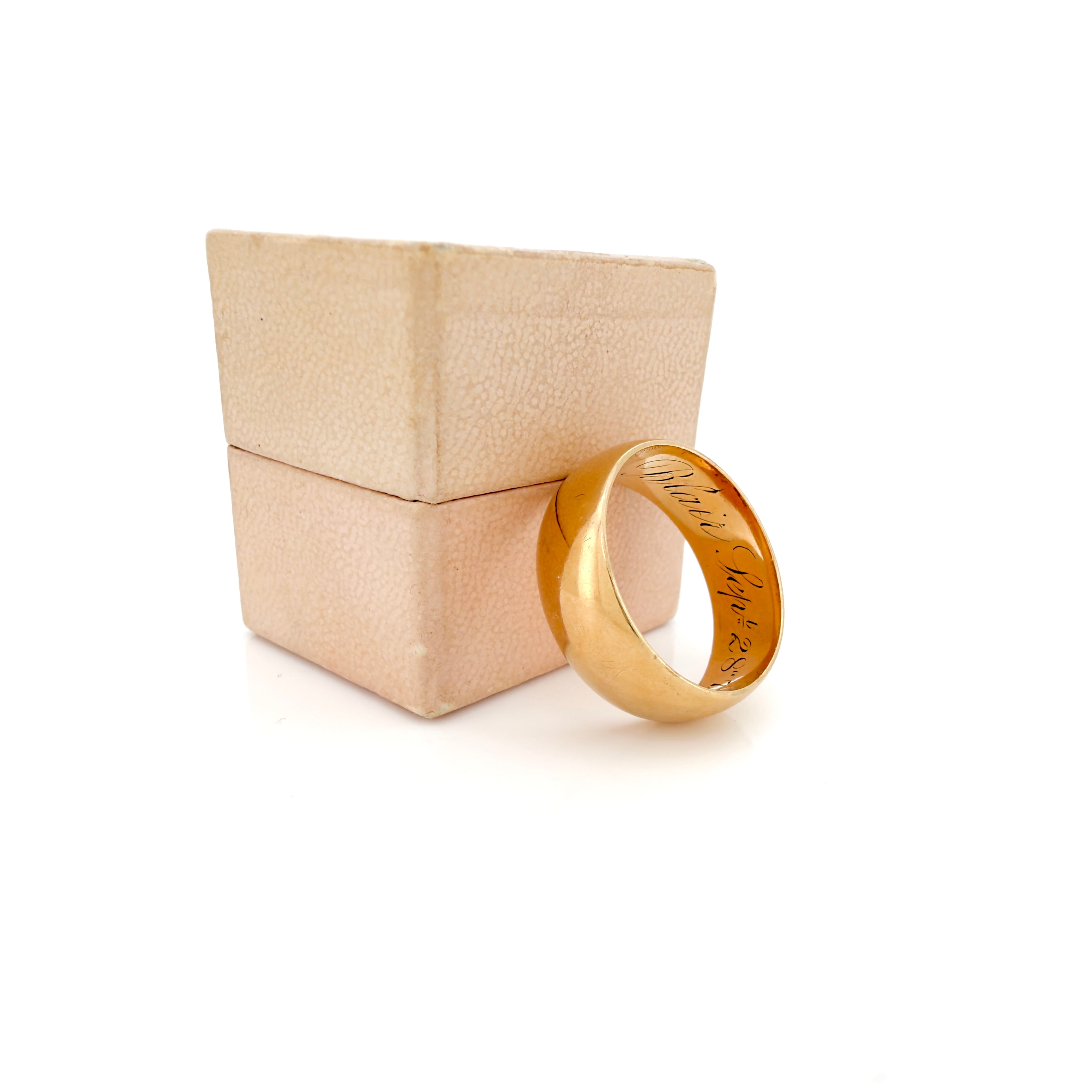 A yellow gold ring with engraved text inside, displayed next to its textured beige presentation box on a white background.