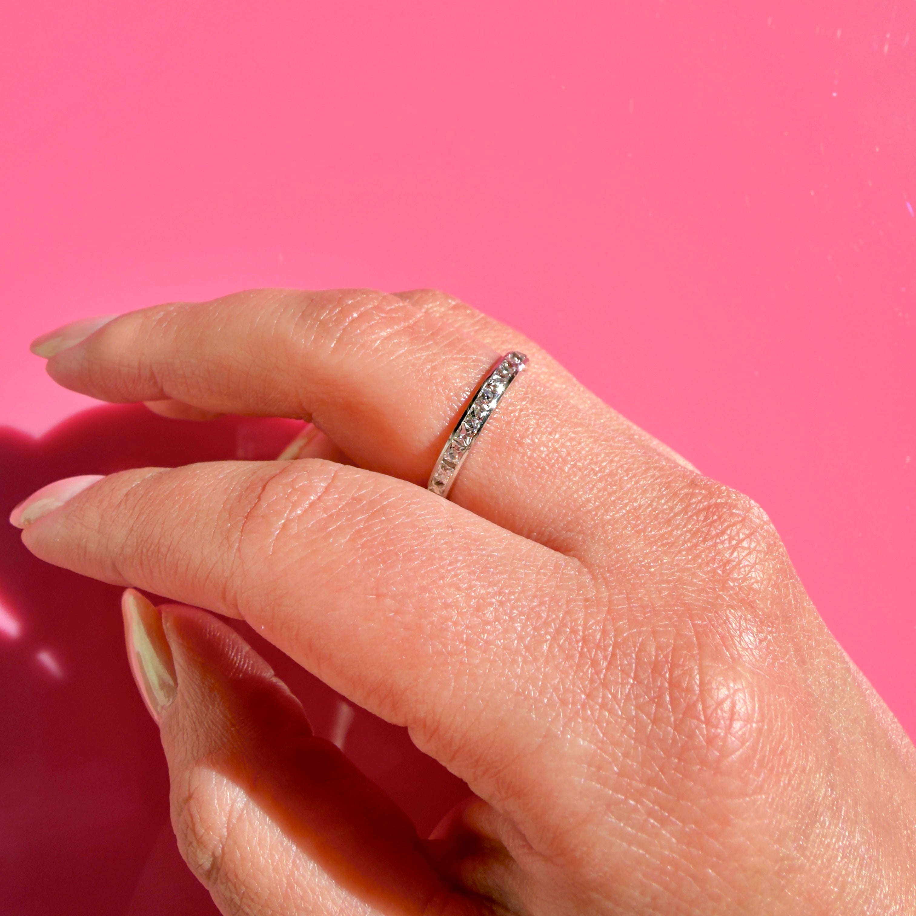A platinum band ring with small French cut diamonds set along the top, worn on a person's ring finger against a solid pink background.