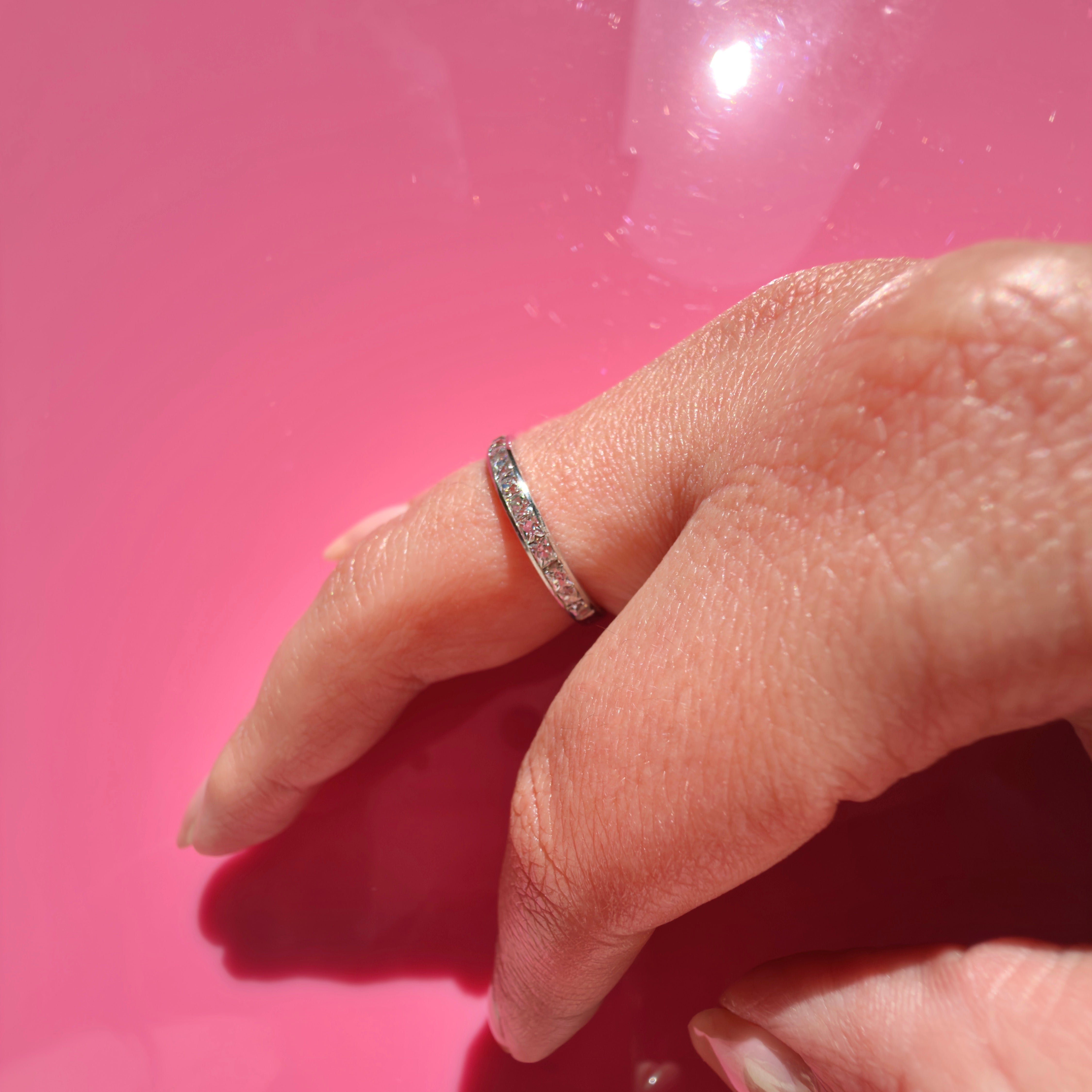 A platinum band ring with small French cut diamonds set along the top, worn on a person's ring finger against a solid pink background.