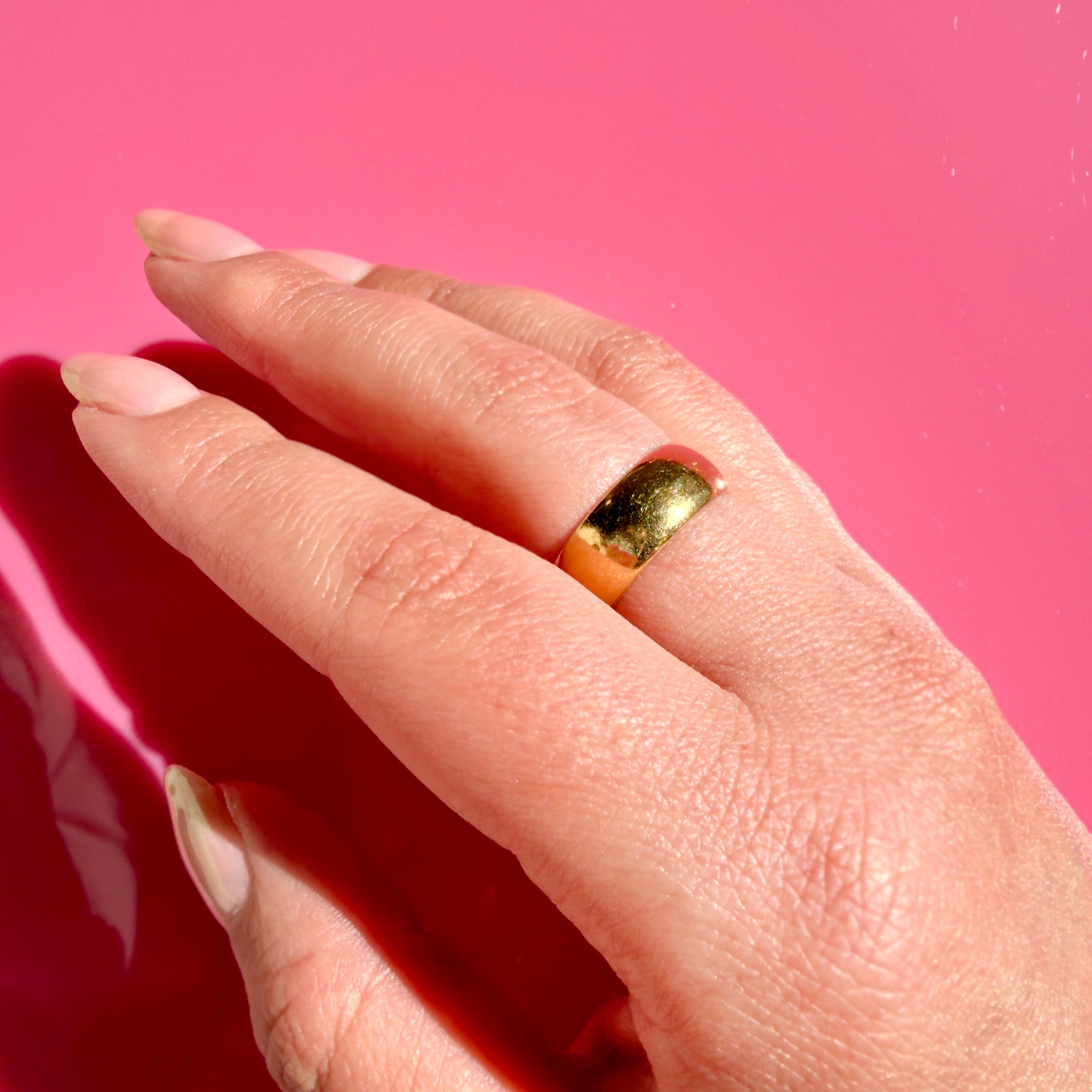 A close-up of a hand wearing a thick, polished gold band ring against a vibrant pink background.