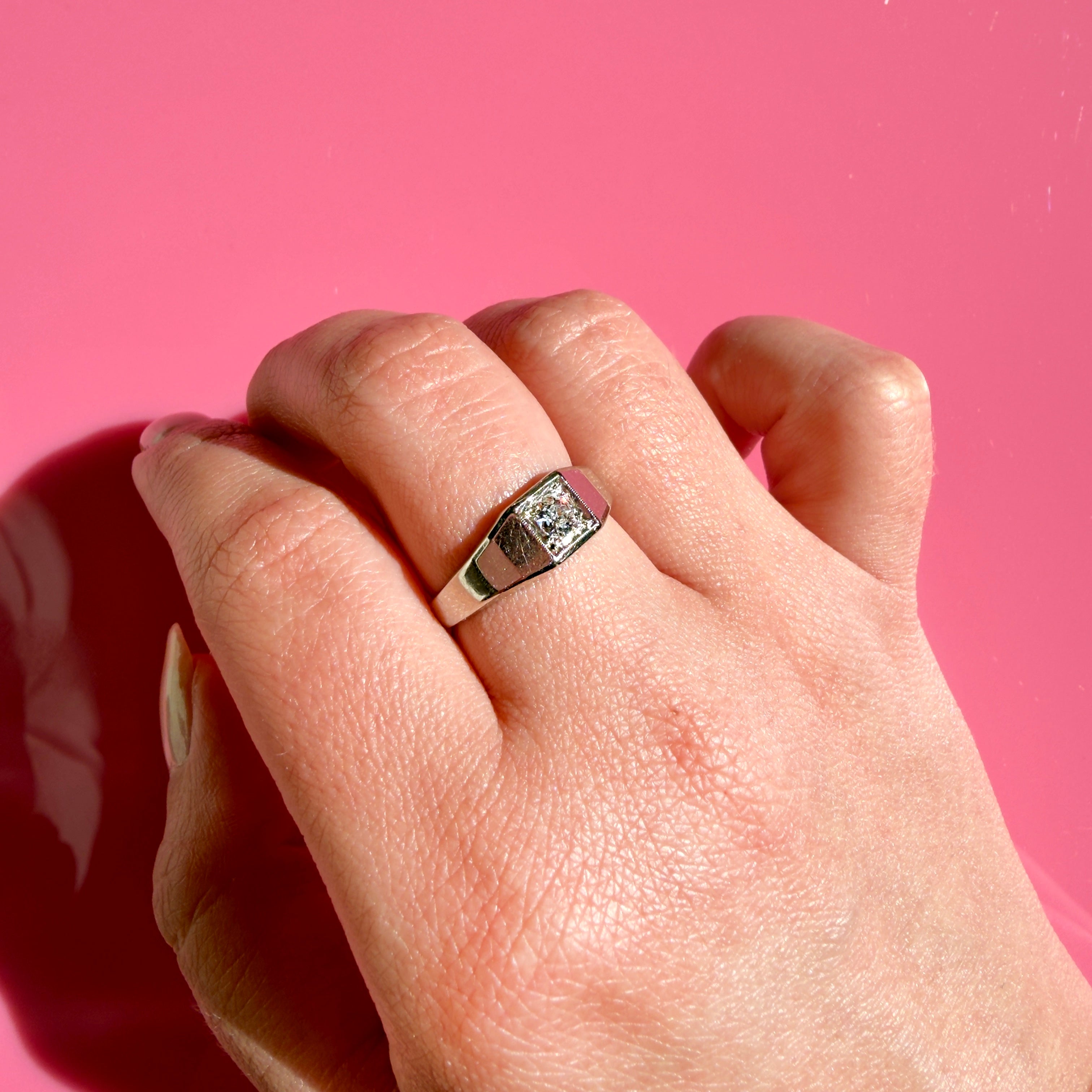 A white gold Art Deco ring with an old European cut diamond set in a geometric bezel-style band, shown worn on a finger against a solid pink background.