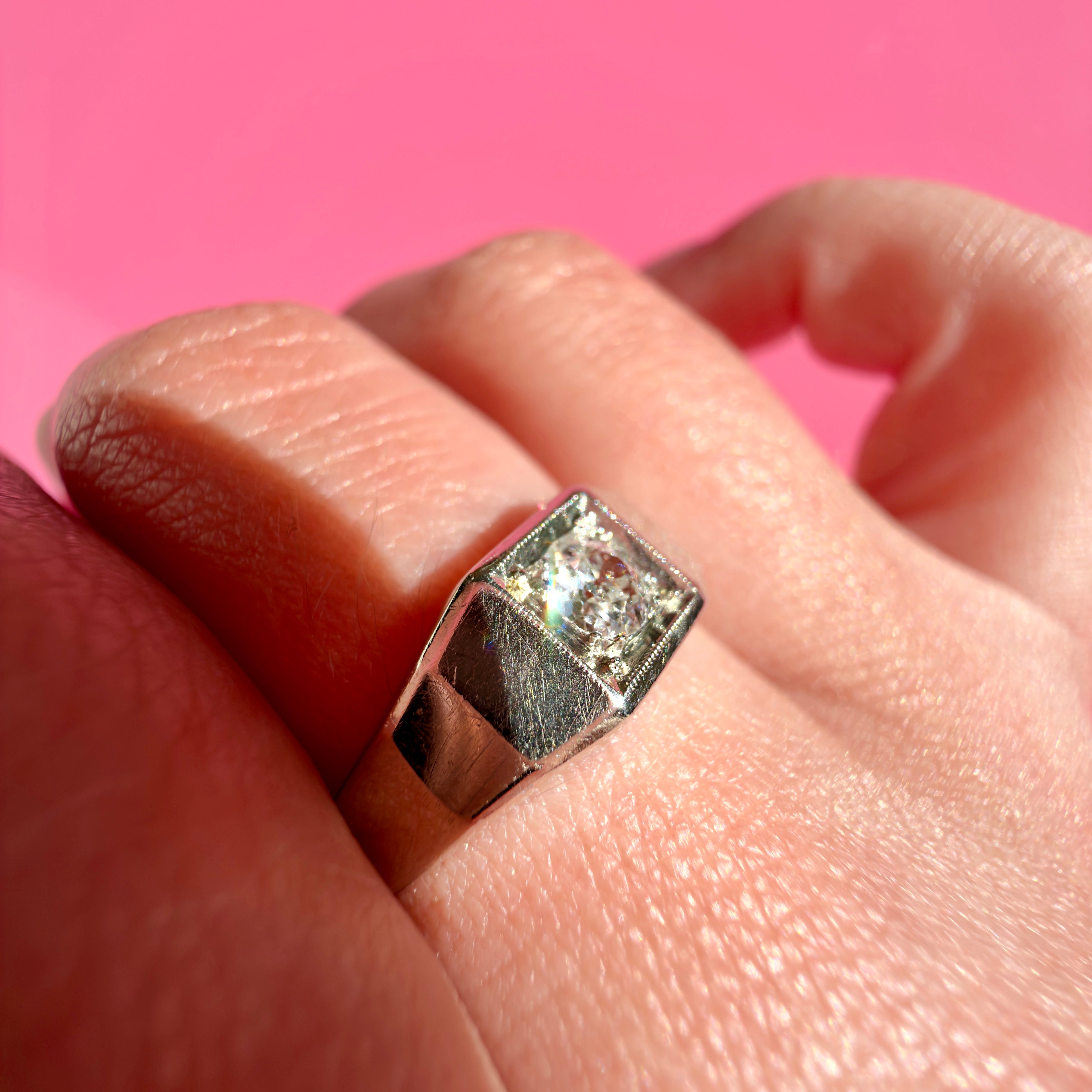A white gold Art Deco ring with an old European cut diamond set in a geometric bezel-style band, shown worn on a finger against a solid pink background.