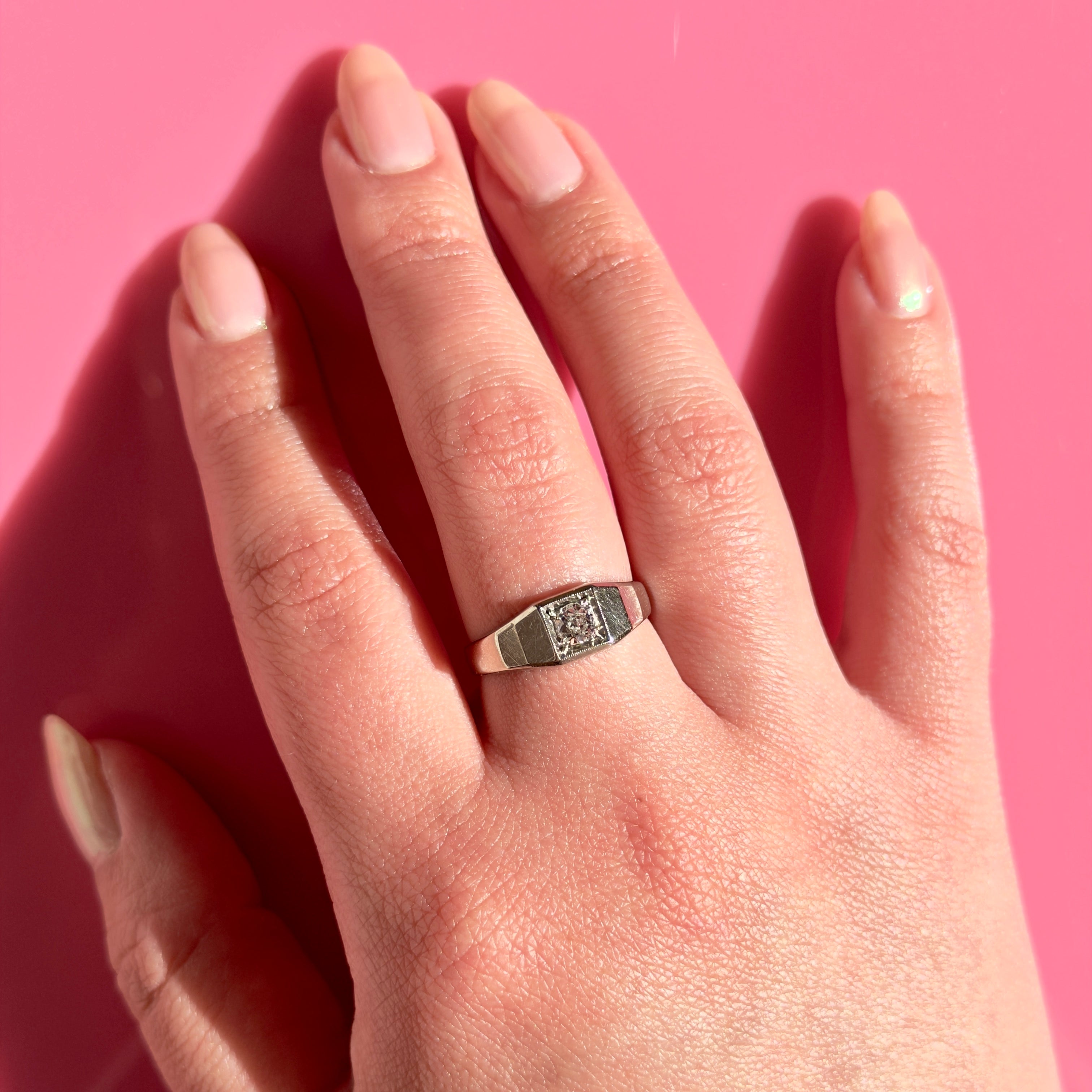 A white gold Art Deco ring with an old European cut diamond set in a geometric bezel-style band, shown worn on a finger against a solid pink background.