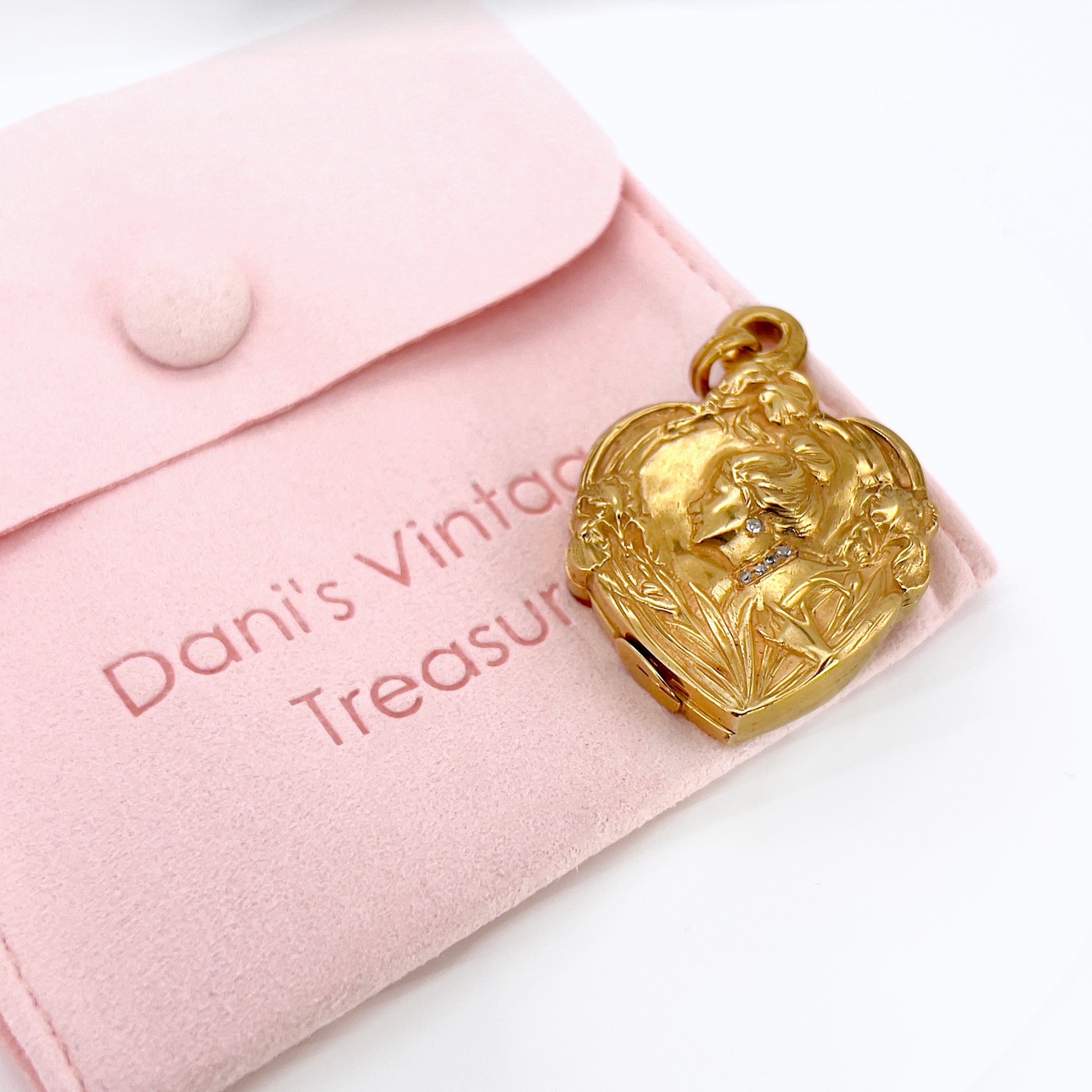An Art Nouveau heart-shaped locket with gold detailing, featuring a central figure with a bun and flowers on the front, on a pink pouch.