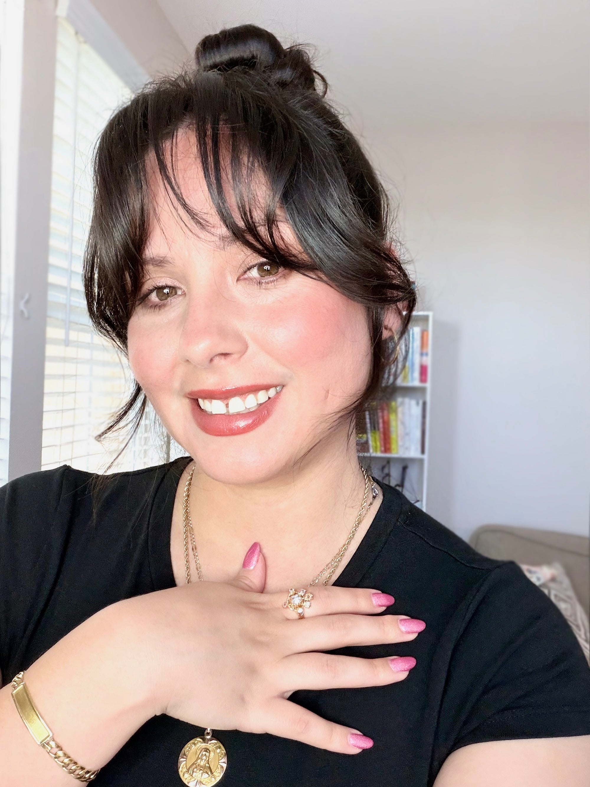 Dani with dark hair in a bun and bangs smiling at the camera, wearing a black t-shirt, layered gold necklaces, a gold bracelet, and a gold ring with pink nail polish, set against an indoor home background.