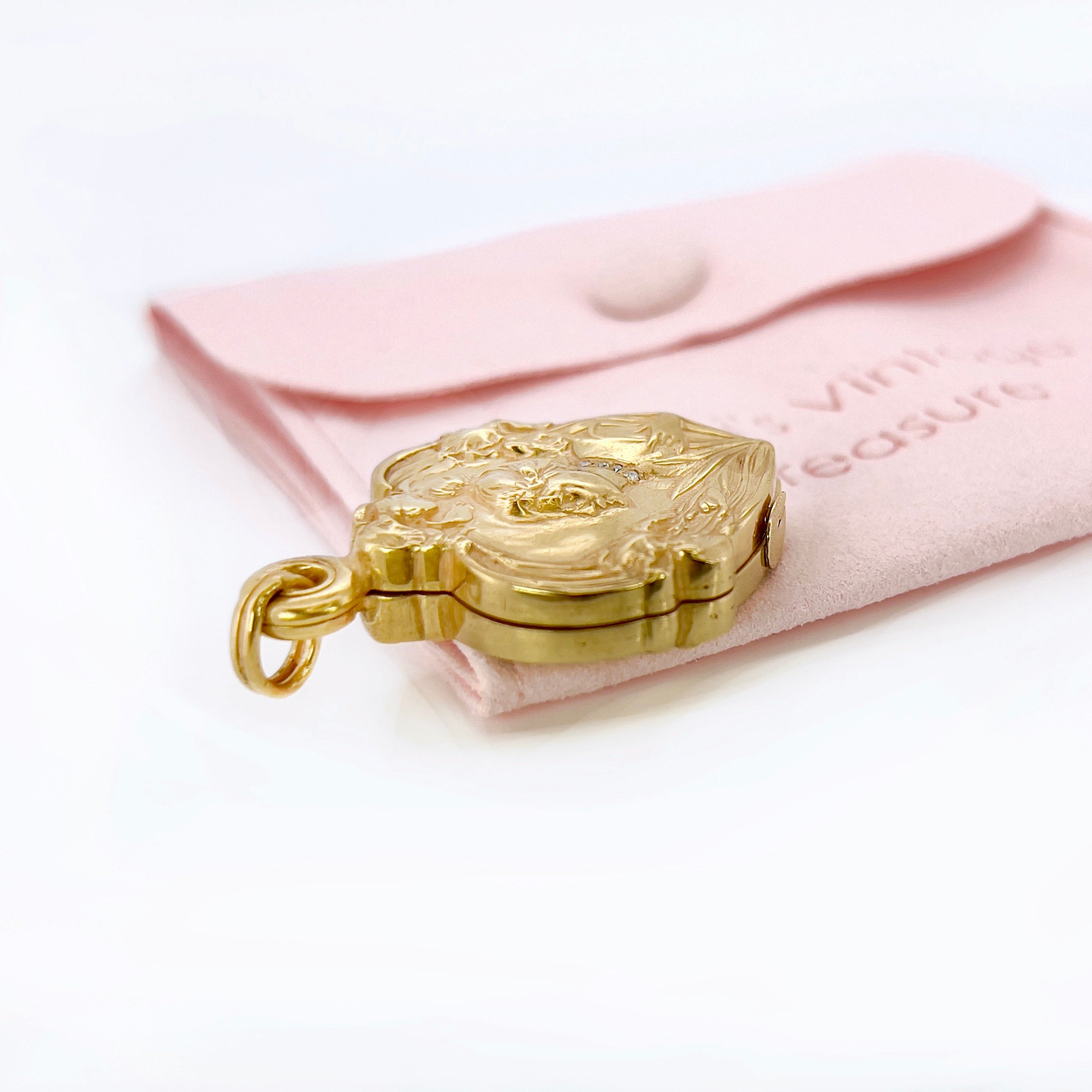 An Art Nouveau heart-shaped locket with gold detailing, featuring a central figure with a bun and flowers on the front, on a pink pouch.