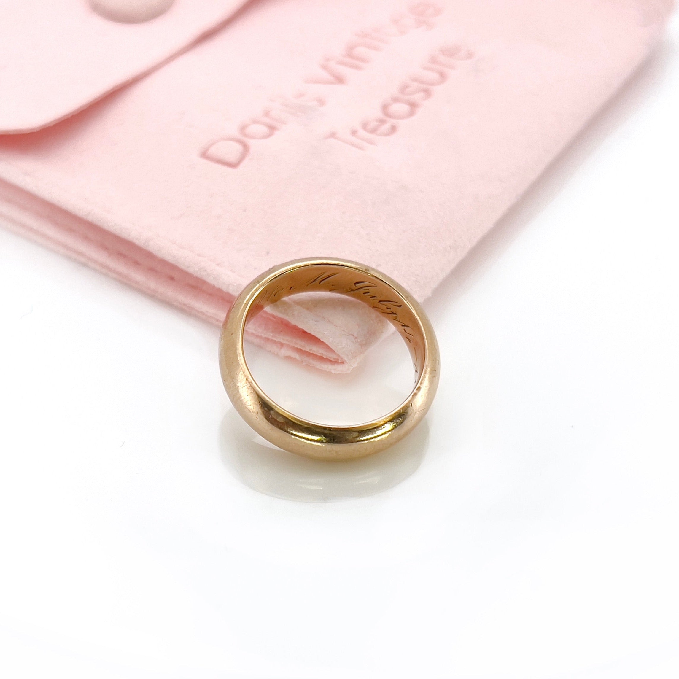 An antique gold band with an inscription, displayed against a pink pouch.