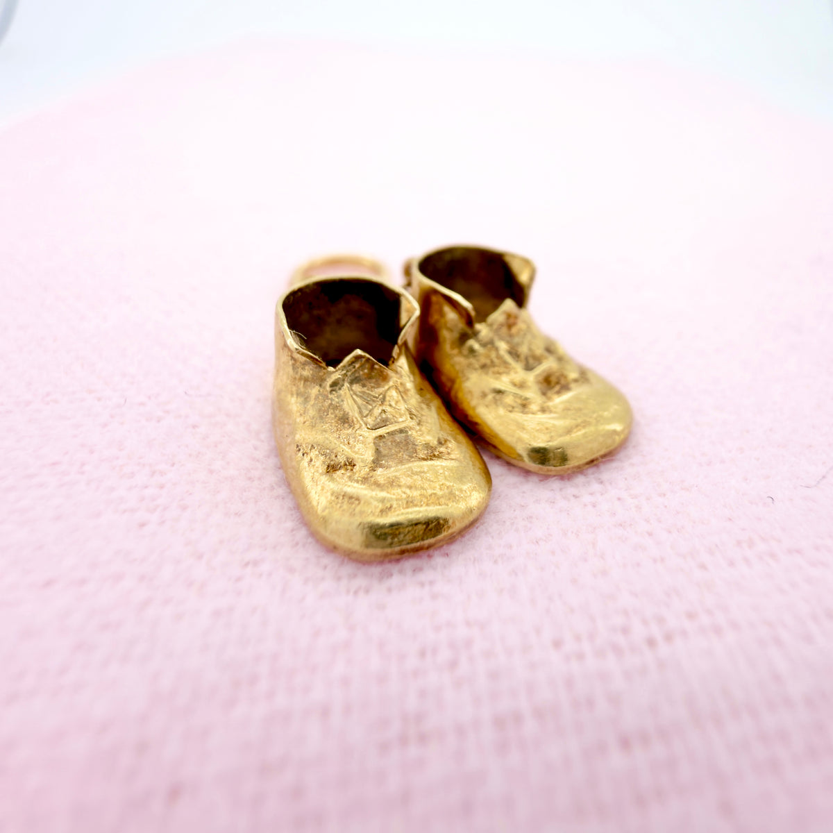 A pair of miniature gold-colored baby shoes, resting on a soft pink textured fabric background.
