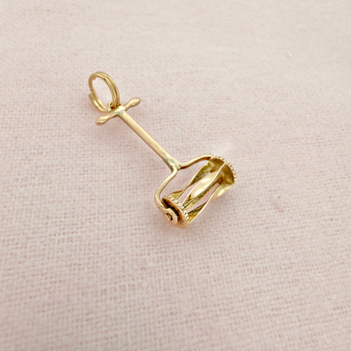Top view of a small gold hand push lawn mower charm with a circular loop at the top, on a textured pink background.