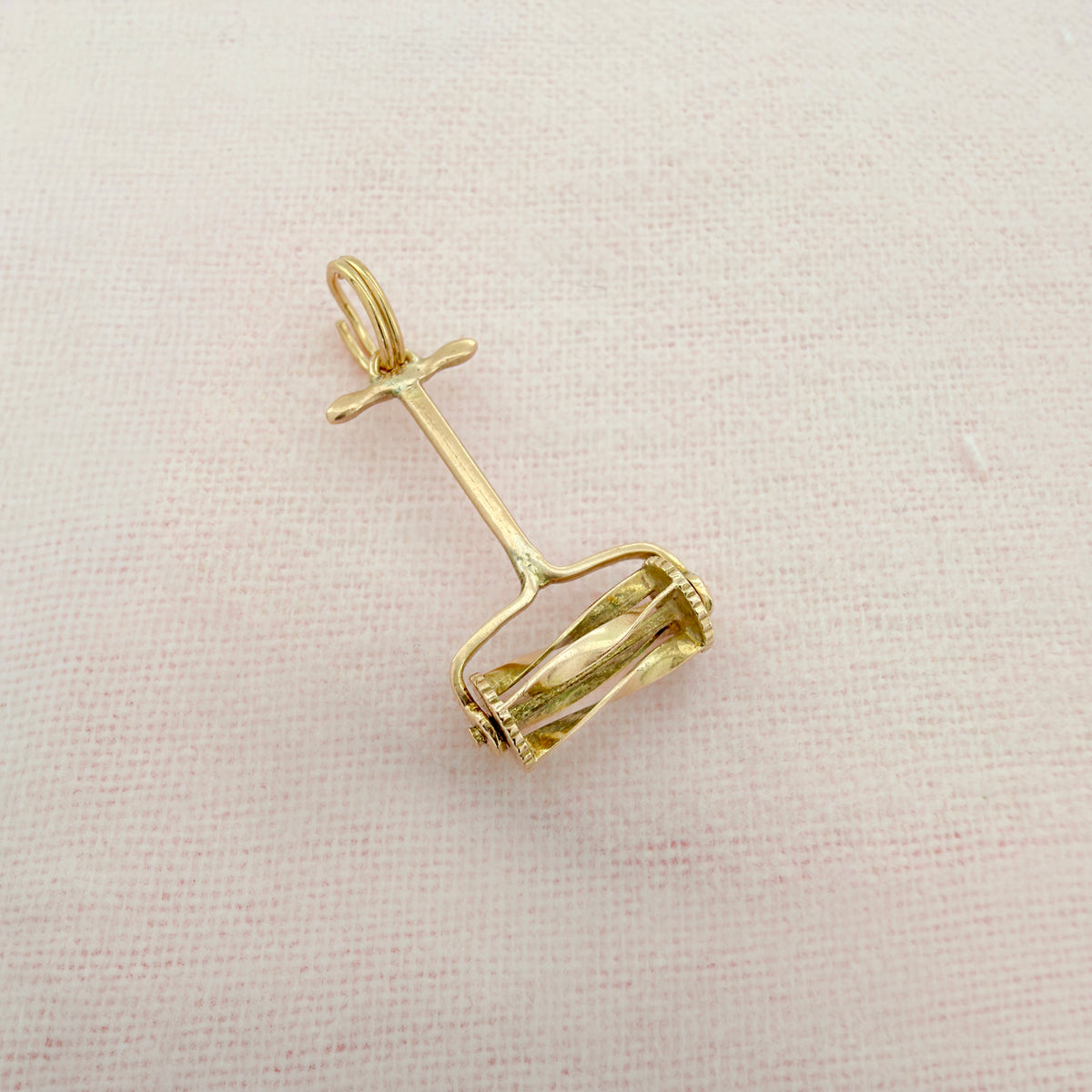 A small gold hand push lawn mower charm with a circular loop at the top and a basket-like openwork lower section, on a textured pink background.