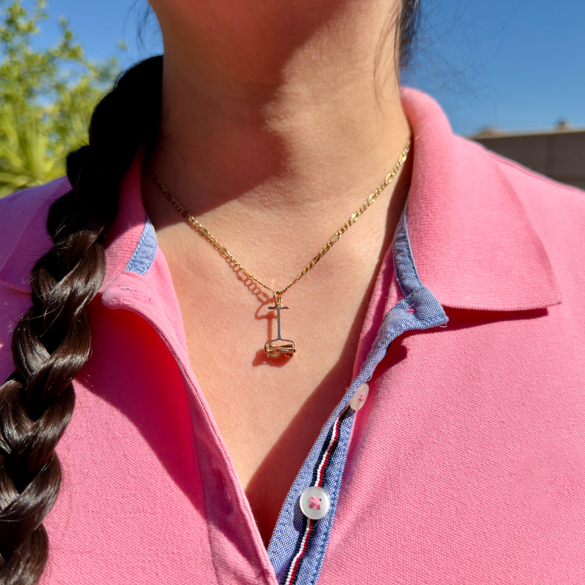 A close-up of a person wearing a delicate gold necklace with a small lawn mower charm, styled with a pink polo shirt and blue collar trim.