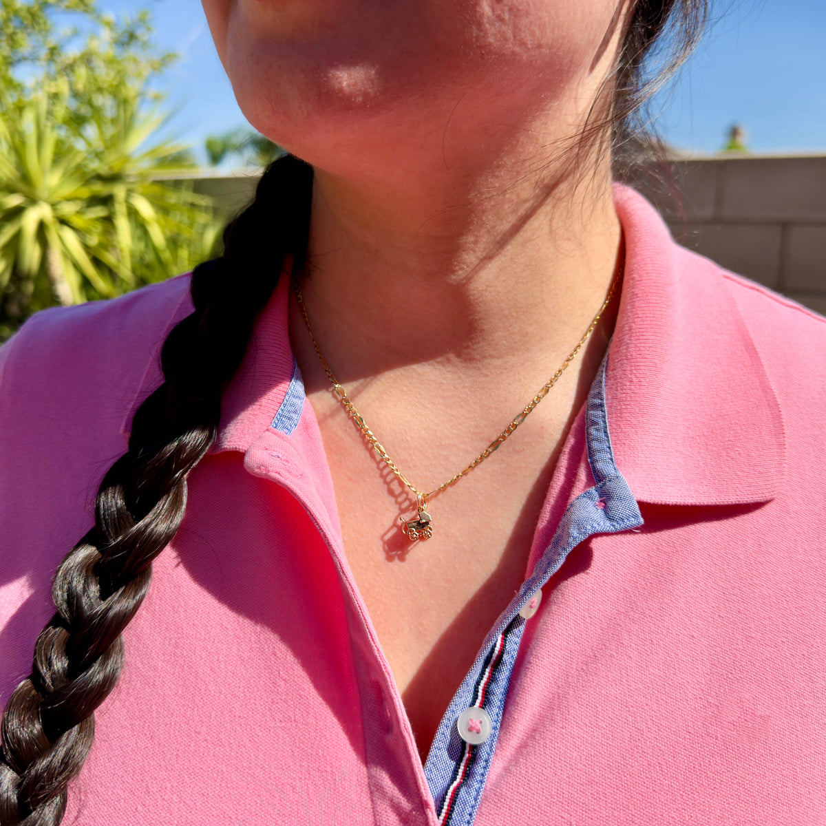 A close-up of a woman wearing a delicate gold chain necklace with a small stroller charm, styled with a pink polo shirt.