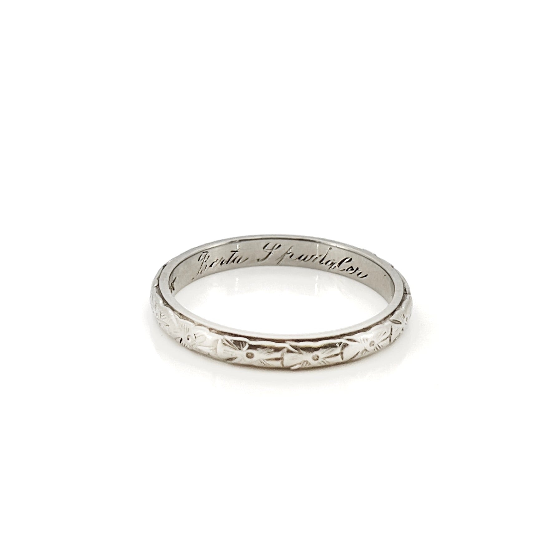 A vintage white gold wedding band with an intricate floral design and the name 'Berta' engraved inside.