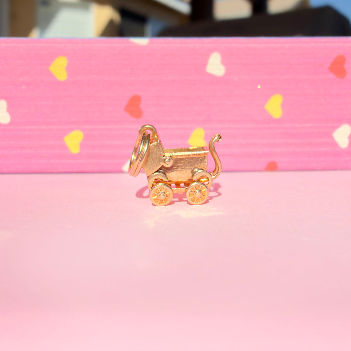 A small gold baby stroller charm with a key ring, displayed on a pink surface with a pink background featuring scattered heart shapes.