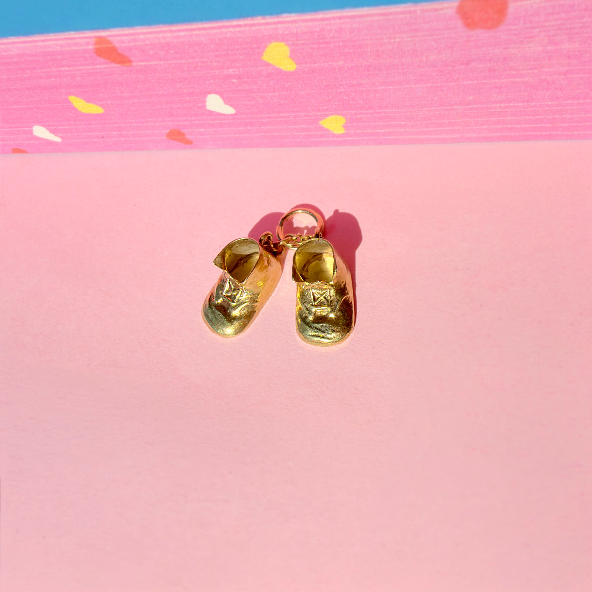 Miniature gold shoe charm with a pink loop at the top, displayed on a pink background with a colorful patterned strip at the top.