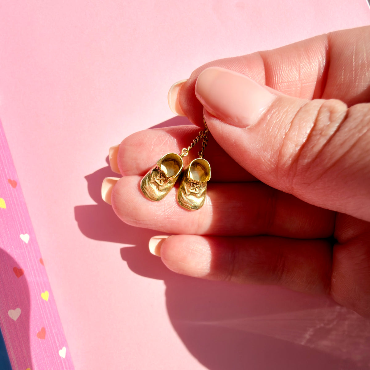 A close-up of a hand holding a pair of tiny gold miniature shoes against a pink background.