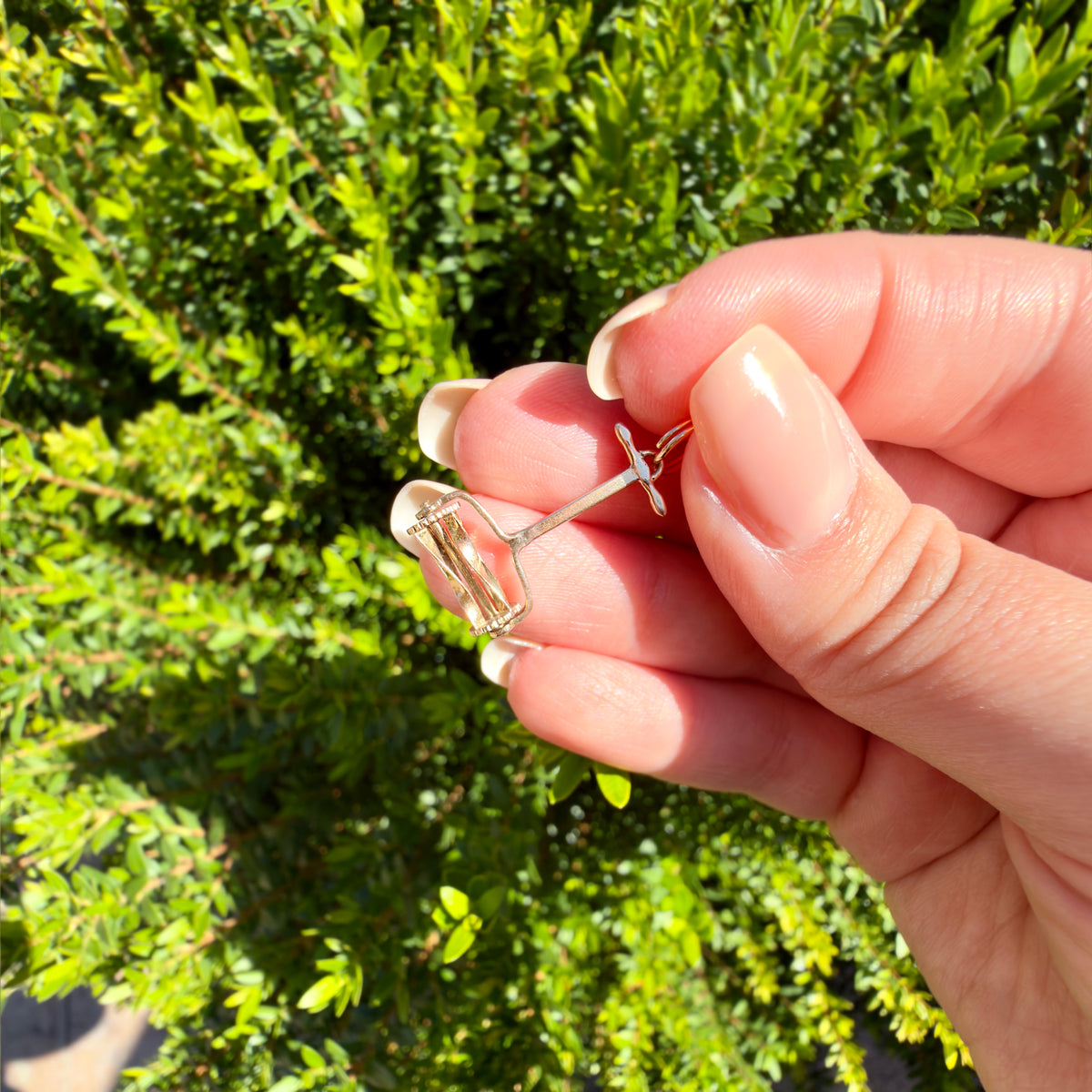 A close-up of a hand holding a small gold lawn mower charm against a blurred green background.