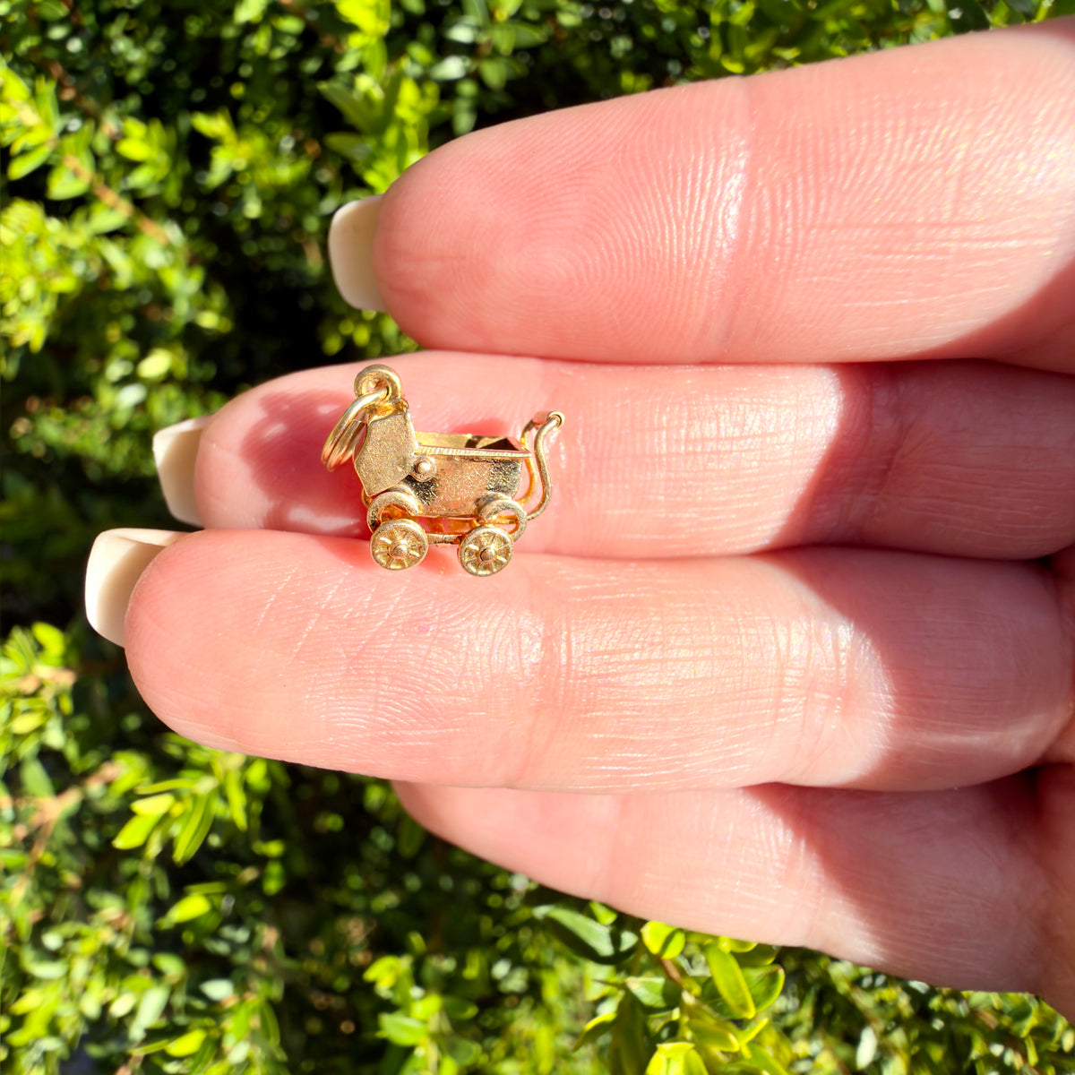 A small gold charm shaped like a stroller resting on a person's fingers, with a blurred green outdoor background.